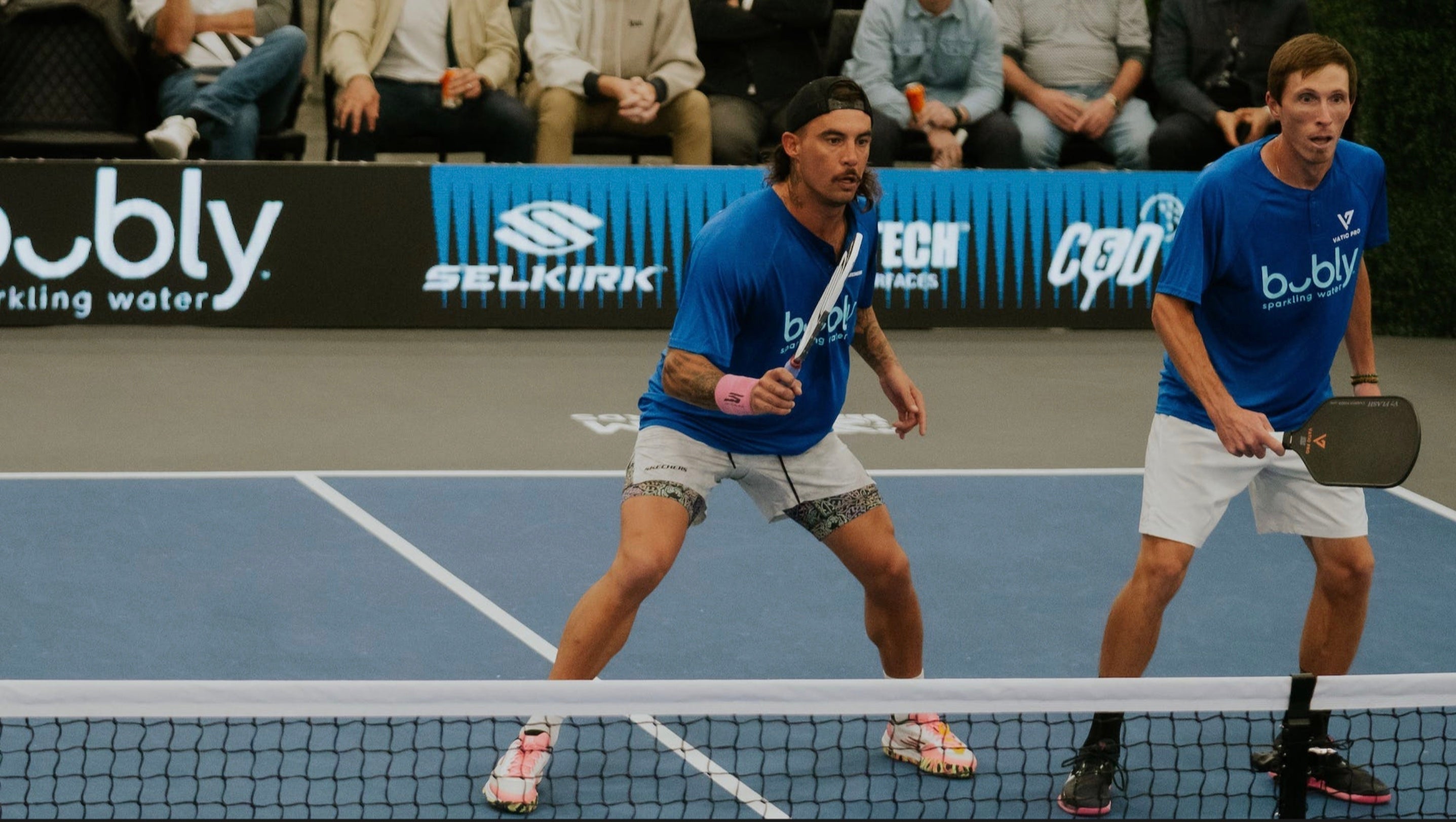 Two pickleball players in blue shirts ready to serve on the court during a tournament.
