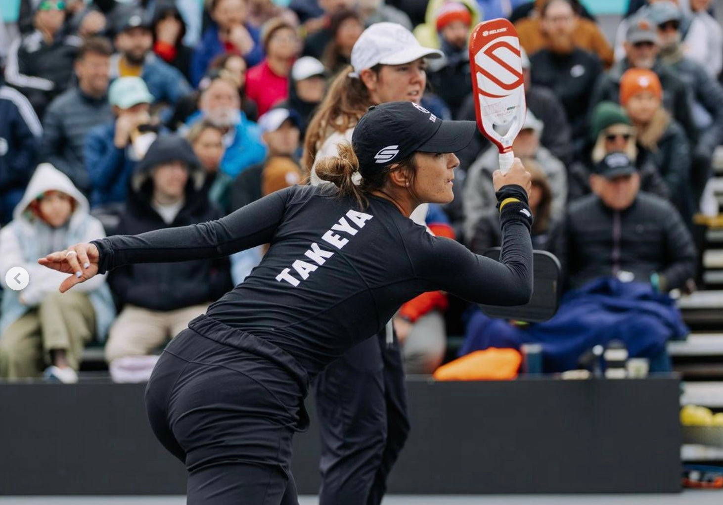 A player in a black outfit with 'TAKEYA' on the back, preparing to hit a pickleball.