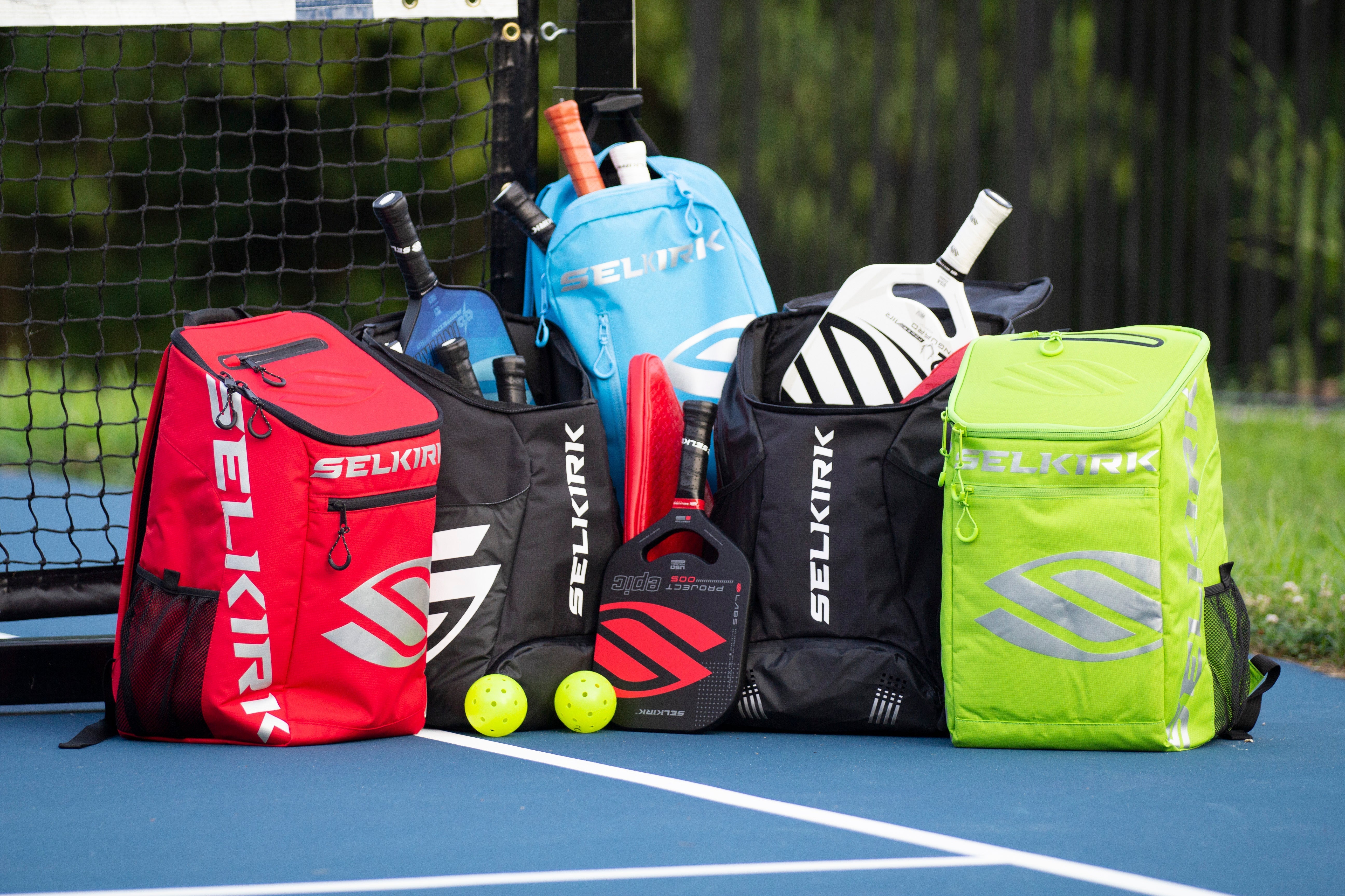 A collection of Selkirk pickleball bags in various colors, including red, blue, black, and green, with paddles and balls visible.