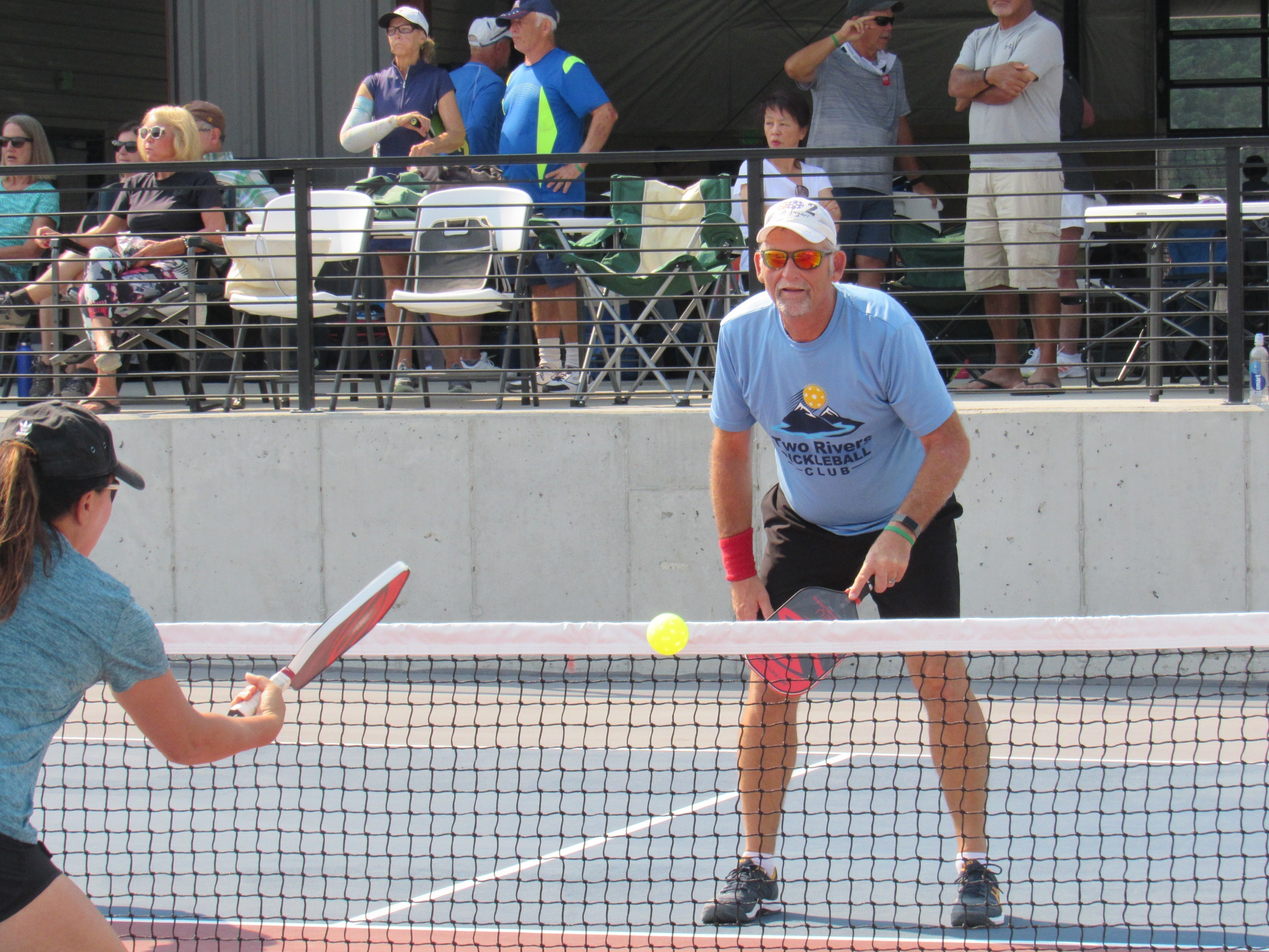 A pickleball match at Two Rivers Pickleball Club, featuring a player preparing to hit the ball at the net.