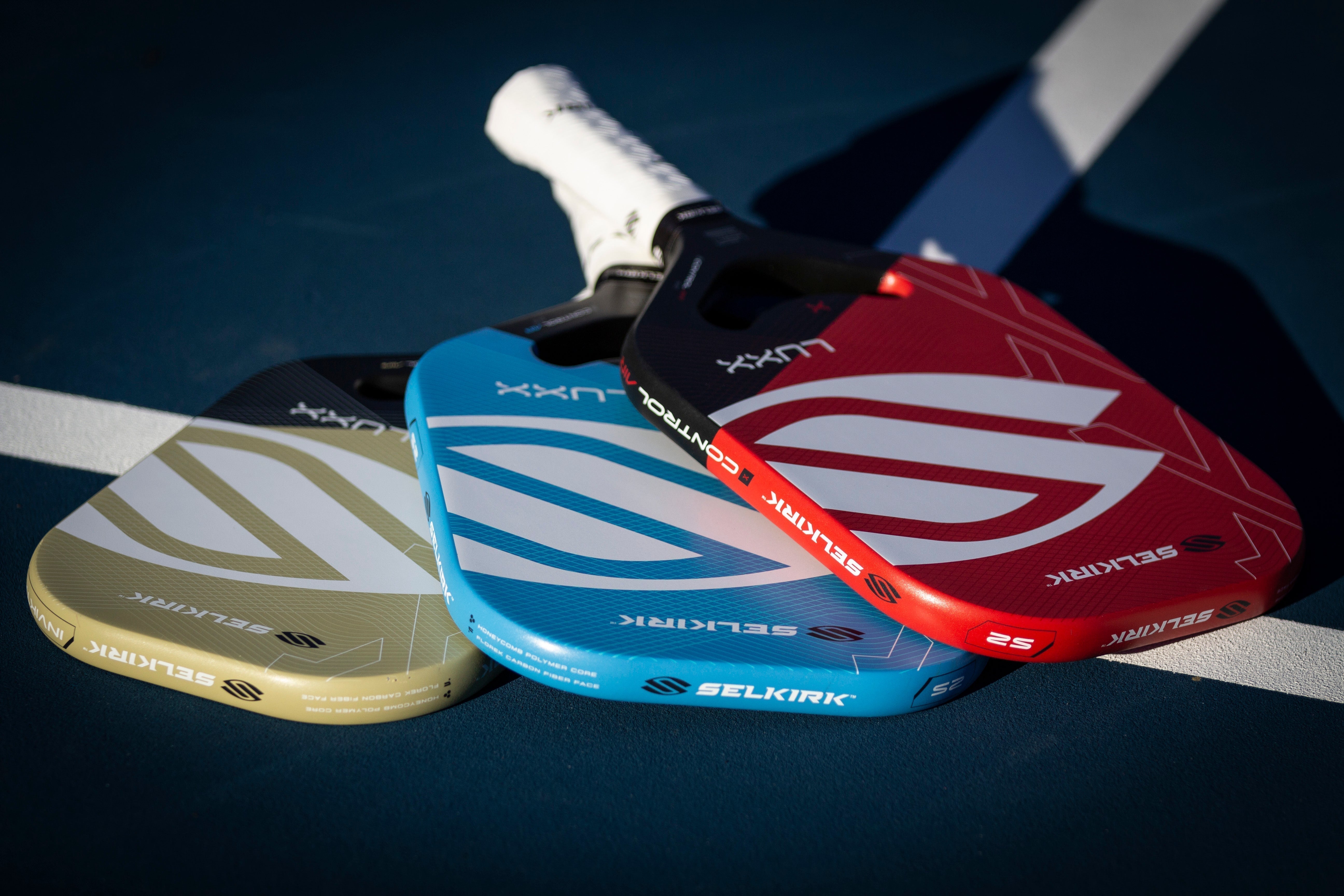 Three Selkirk pickleball paddles in different colors: red, blue, and yellow, displayed on a court.