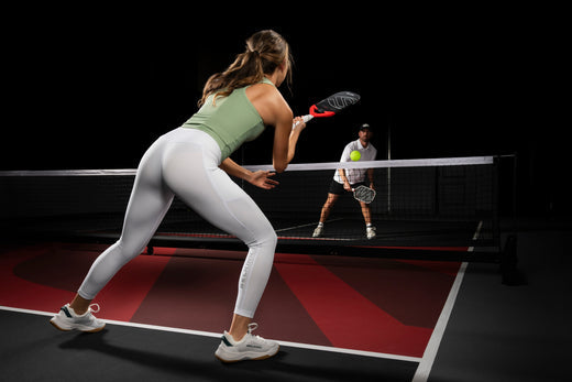 A woman in athletic wear preparing to hit a pickleball, with a man in the background.