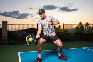 A player in a gray shirt and cap hitting a pickleball with a VANGUARD Pro paddle on a court.
