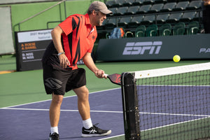 A man in a red and black shirt playing pickleball, preparing to hit a ball over the net.