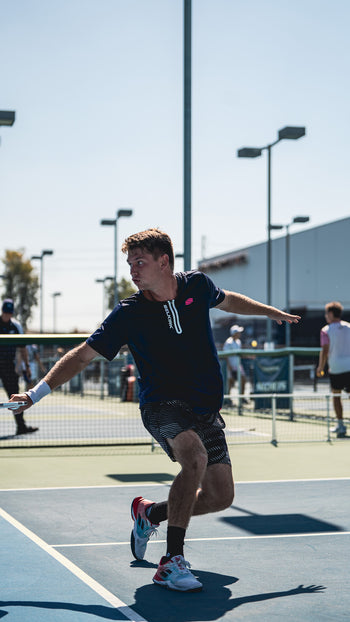Young player lunging to return a shot on an outdoor tennis court, shadow cast on the court surface.