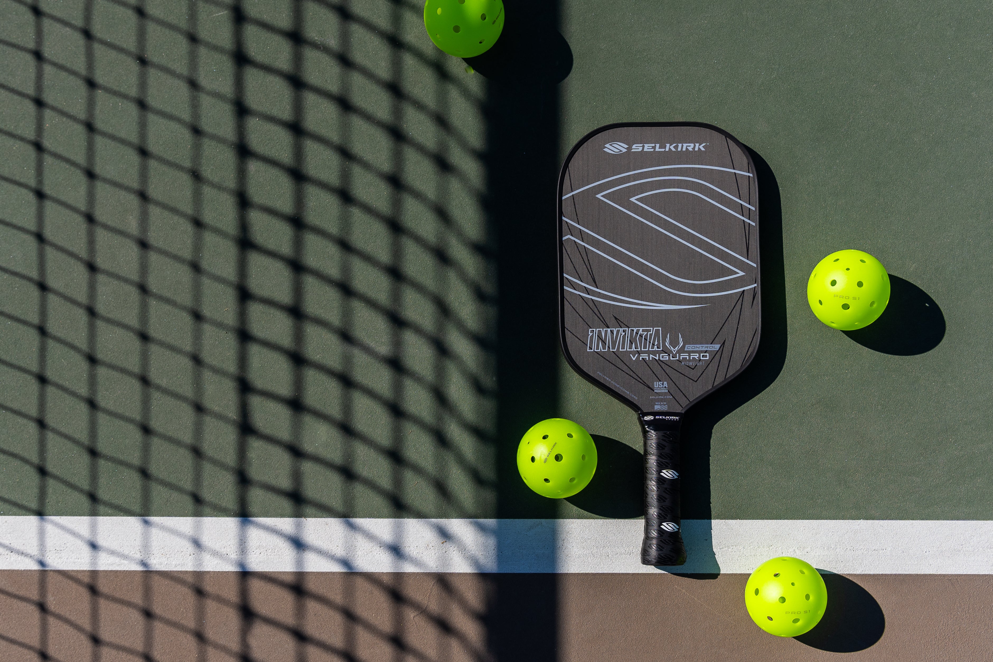 Selkirk Vanguard Invikta lying on a pickleball court with pickleball balls surrounding it.