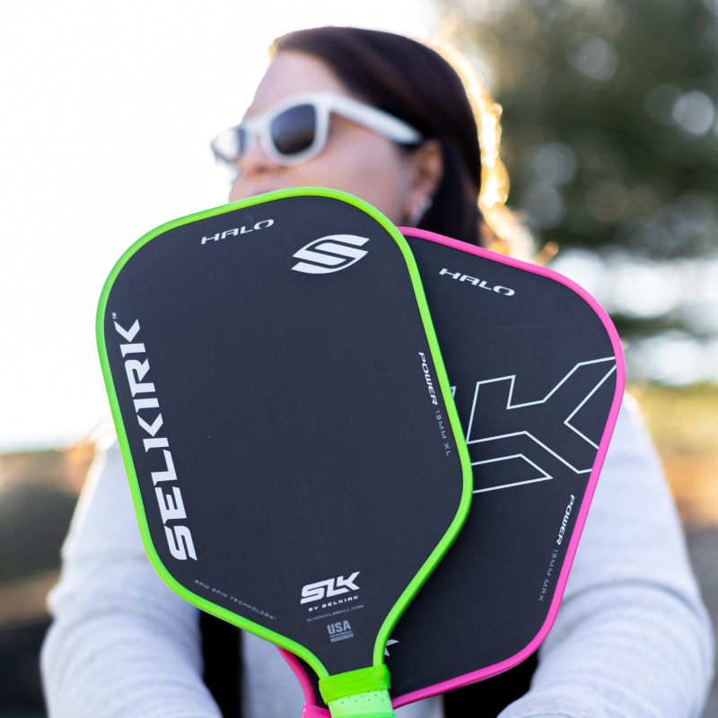Woman holding up 2 pickleball paddles, one with a green edgeguard and the other with a pink edgeguard.