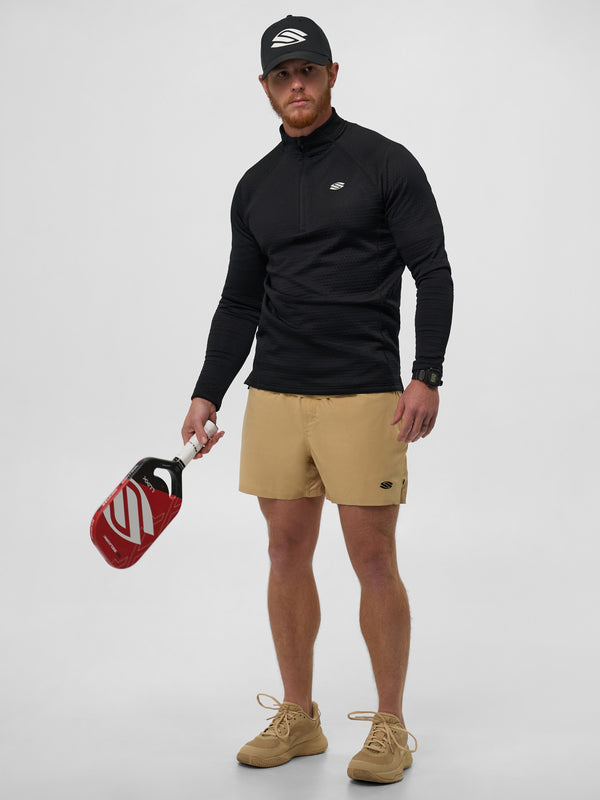Model wearing a black 1/4 zip pullover and beige shorts, holding a pickleball paddle.