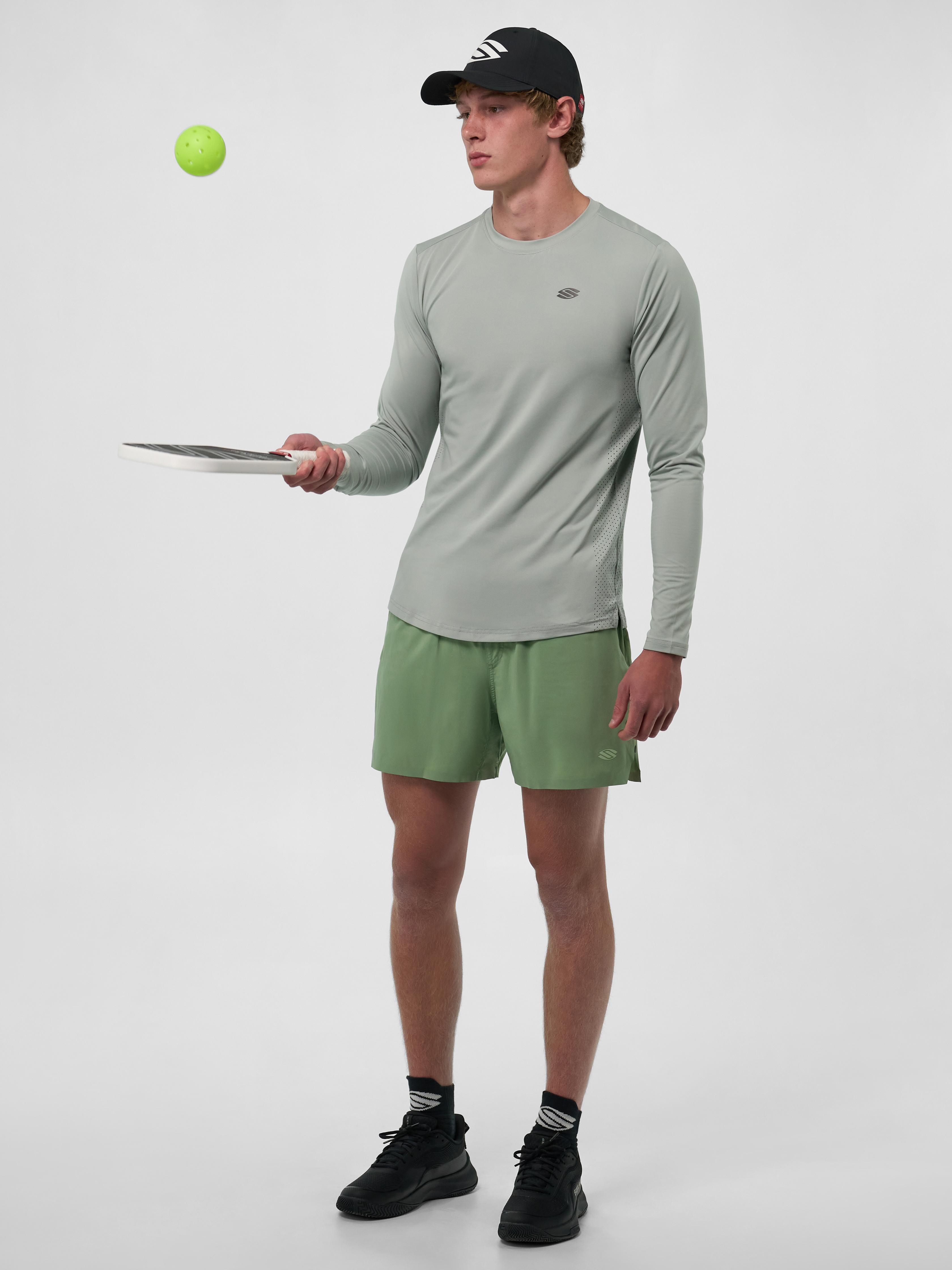 Model wearing a light green long sleeve athletic shirt and green shorts, holding a pickleball paddle with a ball in the air.