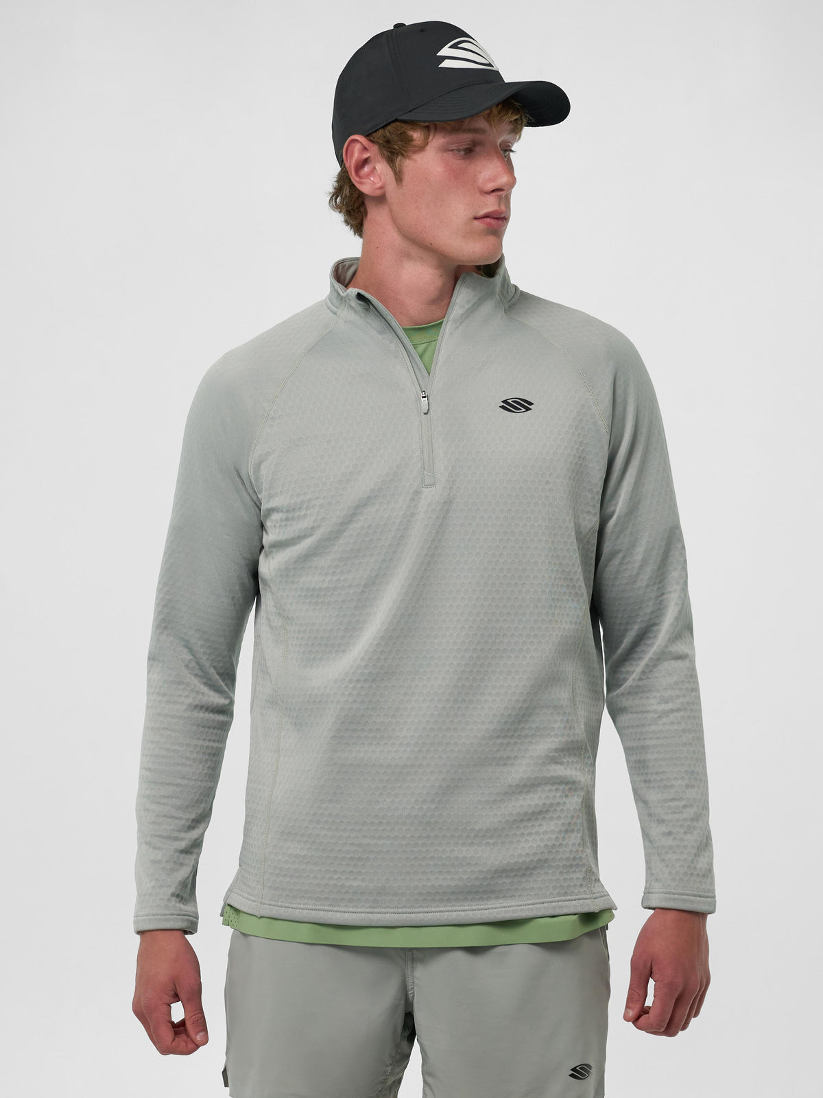 Model wearing a Selkirk men's quarter-zip shirt in light grey, with a black cap, standing against a neutral background.