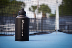 A Selkirk water bottle stands upright near a pickleball net. 