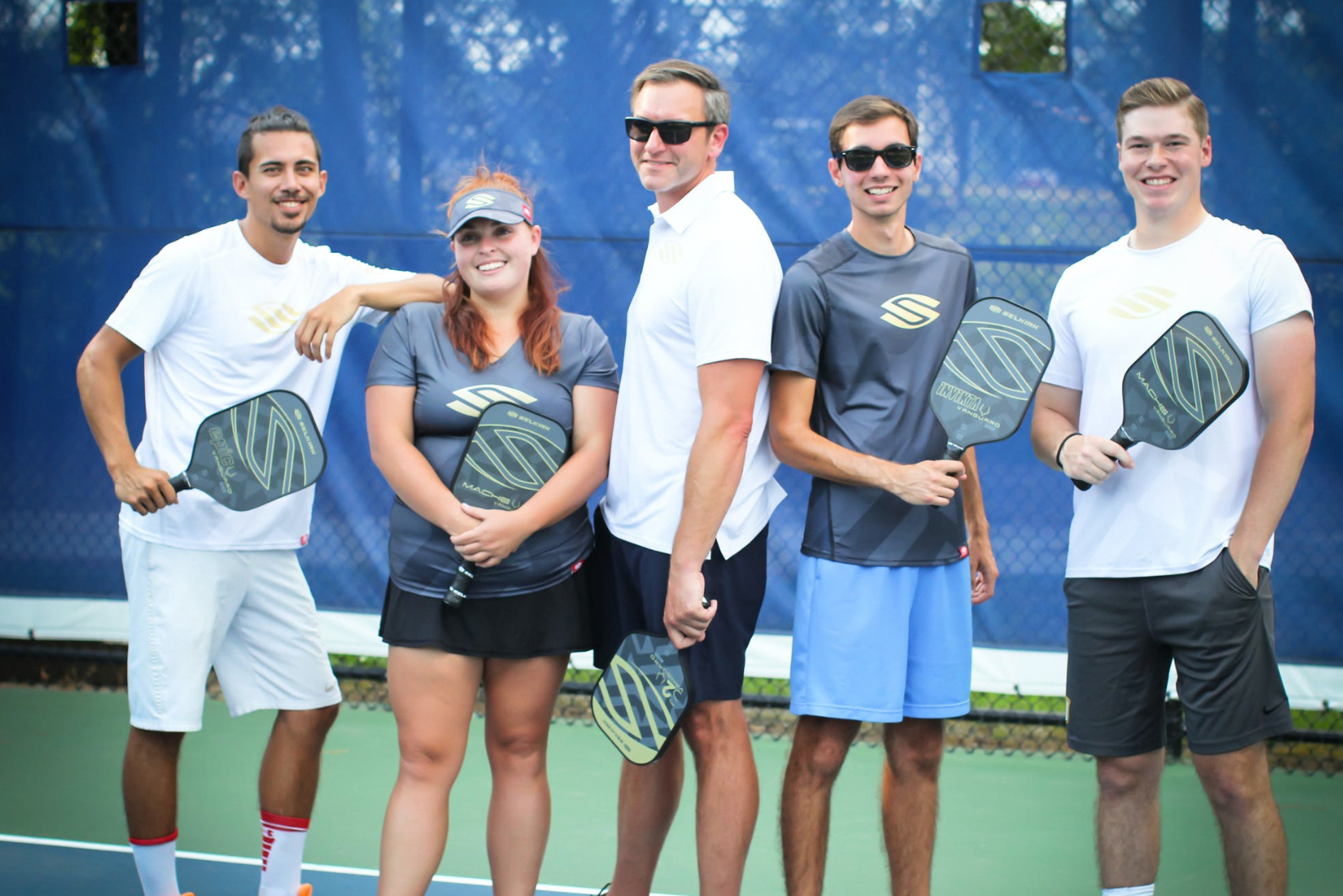 Five players posing with pickleball paddles on a court