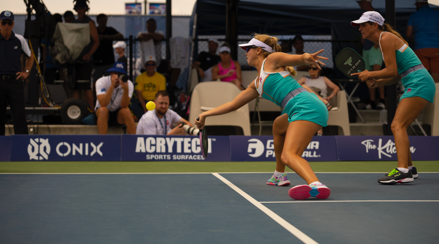 A player lunges to hit a pickleball while another player prepares to receive it during a tournament match.