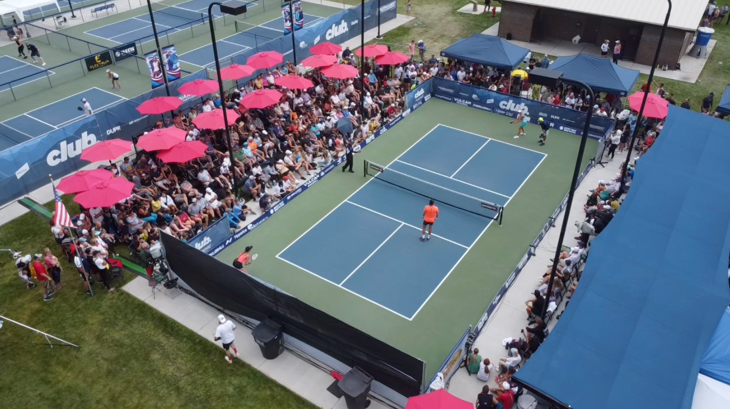 Aerial view of a crowded pickleball court with players and spectators under red umbrellas.