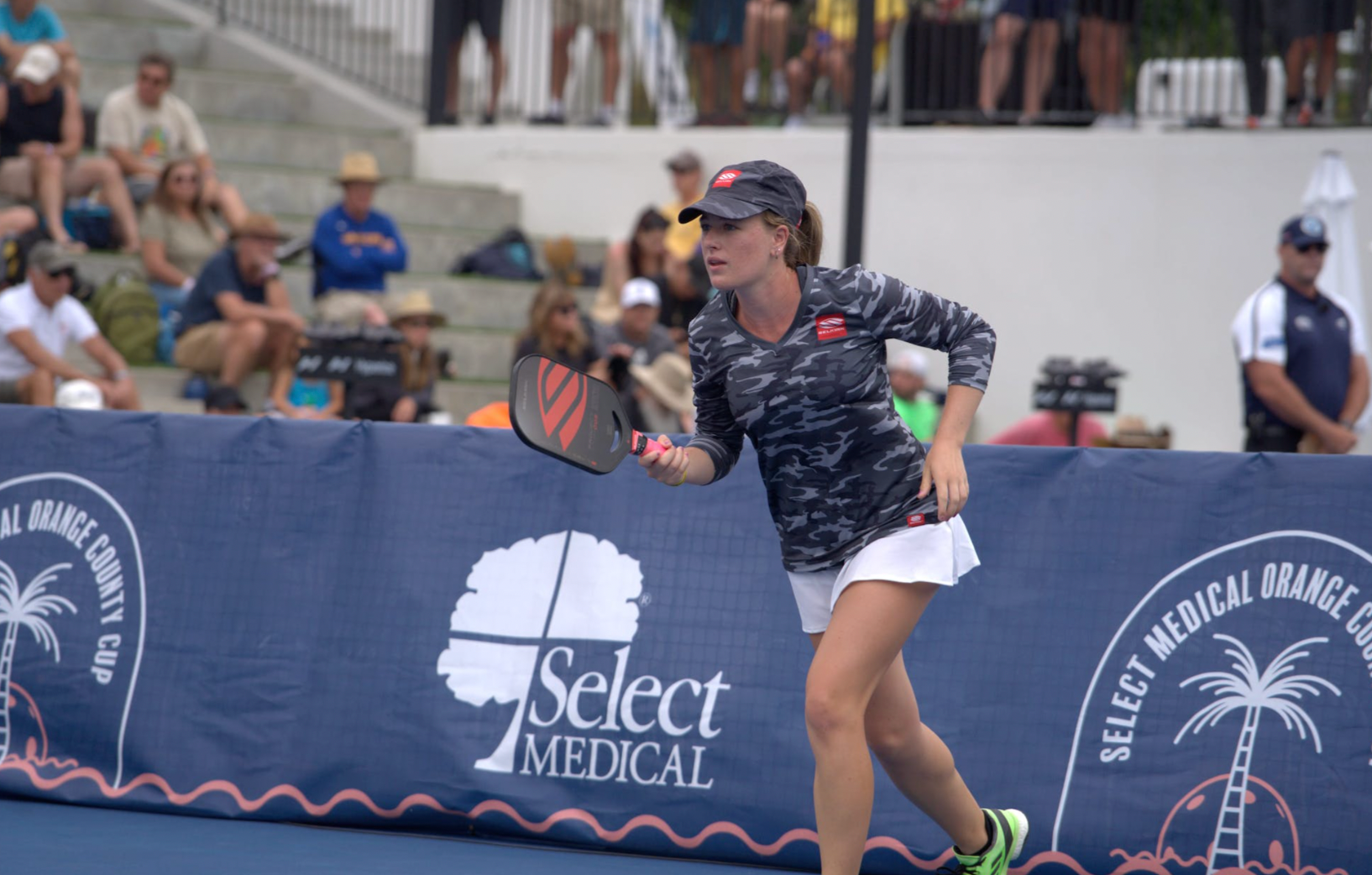 A female pickleball player in action at the Select Medical Orange County Cup, with a banner displaying 'Select Medical'.