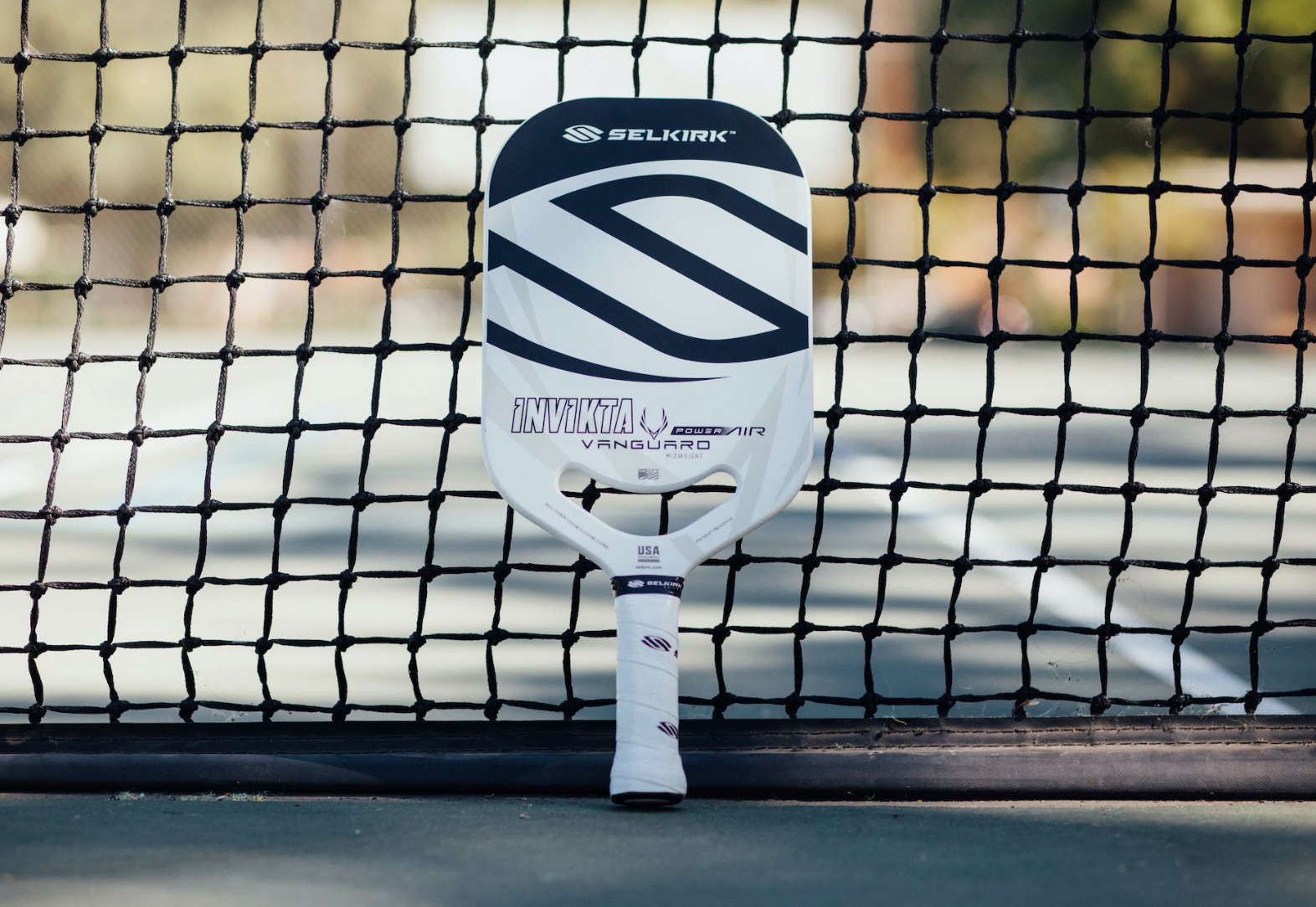 The VANGUARD Power Air paddle, Invikta shape, leaning against a pickleball net.
