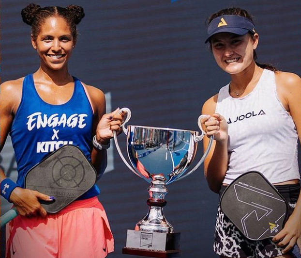 Two women holding a trophy, one in a blue shirt and the other in a white shirt, celebrating their victory.
