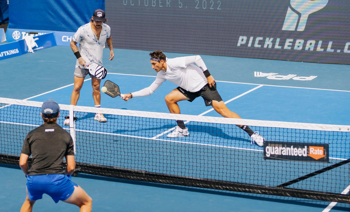 Two players competing in a pickleball match on a blue court, with one player reaching for the ball.