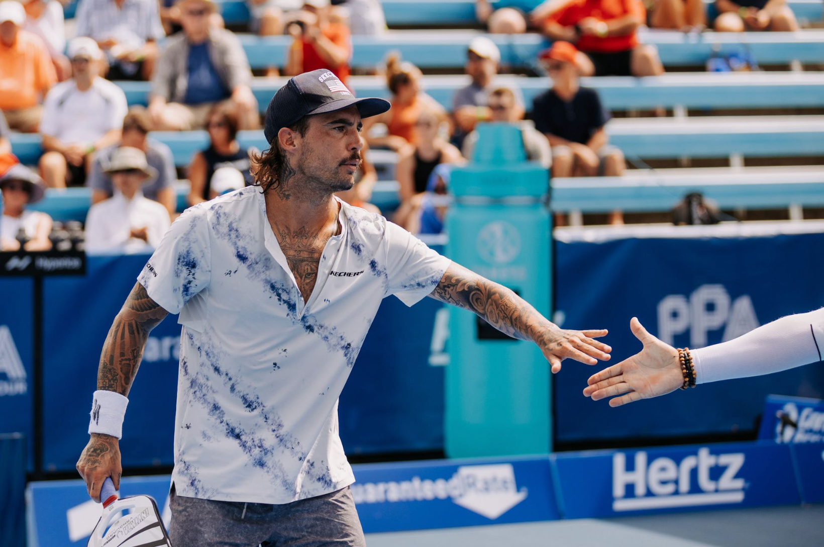 A man in a blue shirt and cap reaching out for a handshake on a pickleball court.