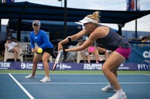 Two women play Pickleball on an outdoor court while spectators watch