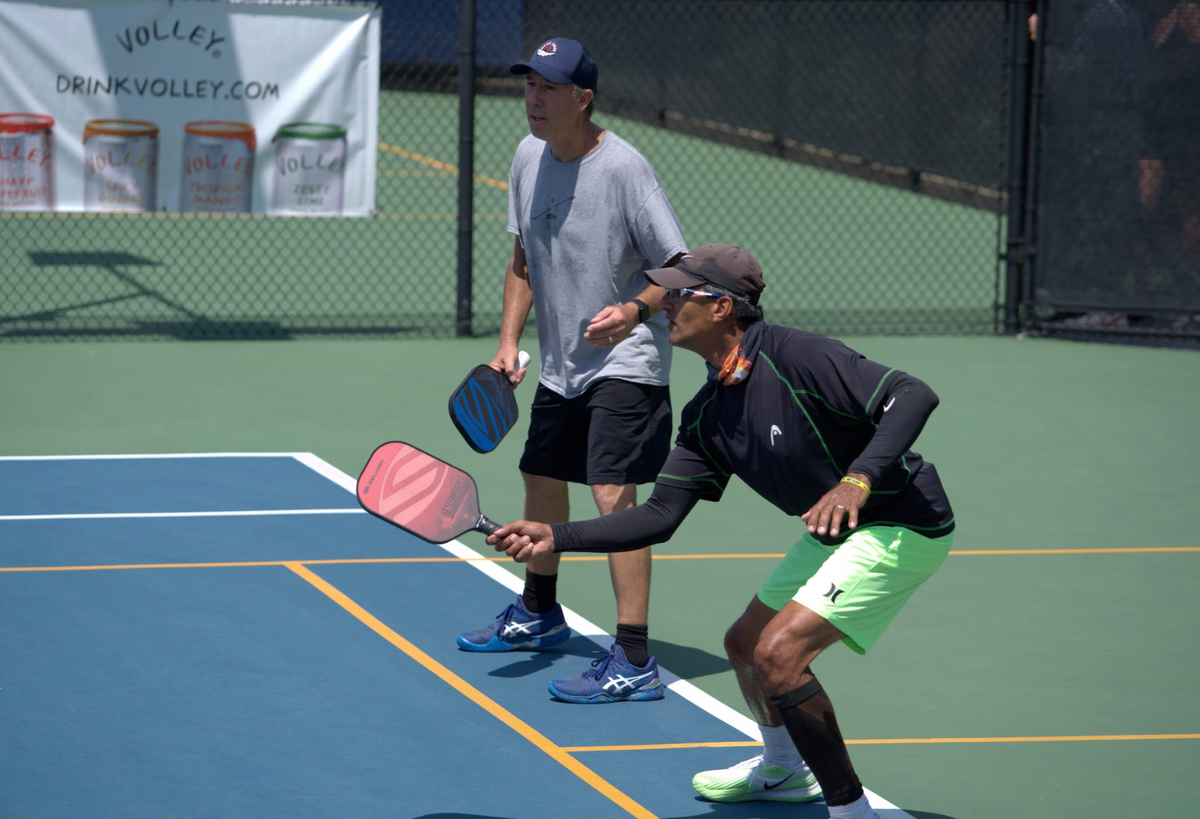 Two men play pickleball on an outdoor court, one practicing a forehand volley while the other observes