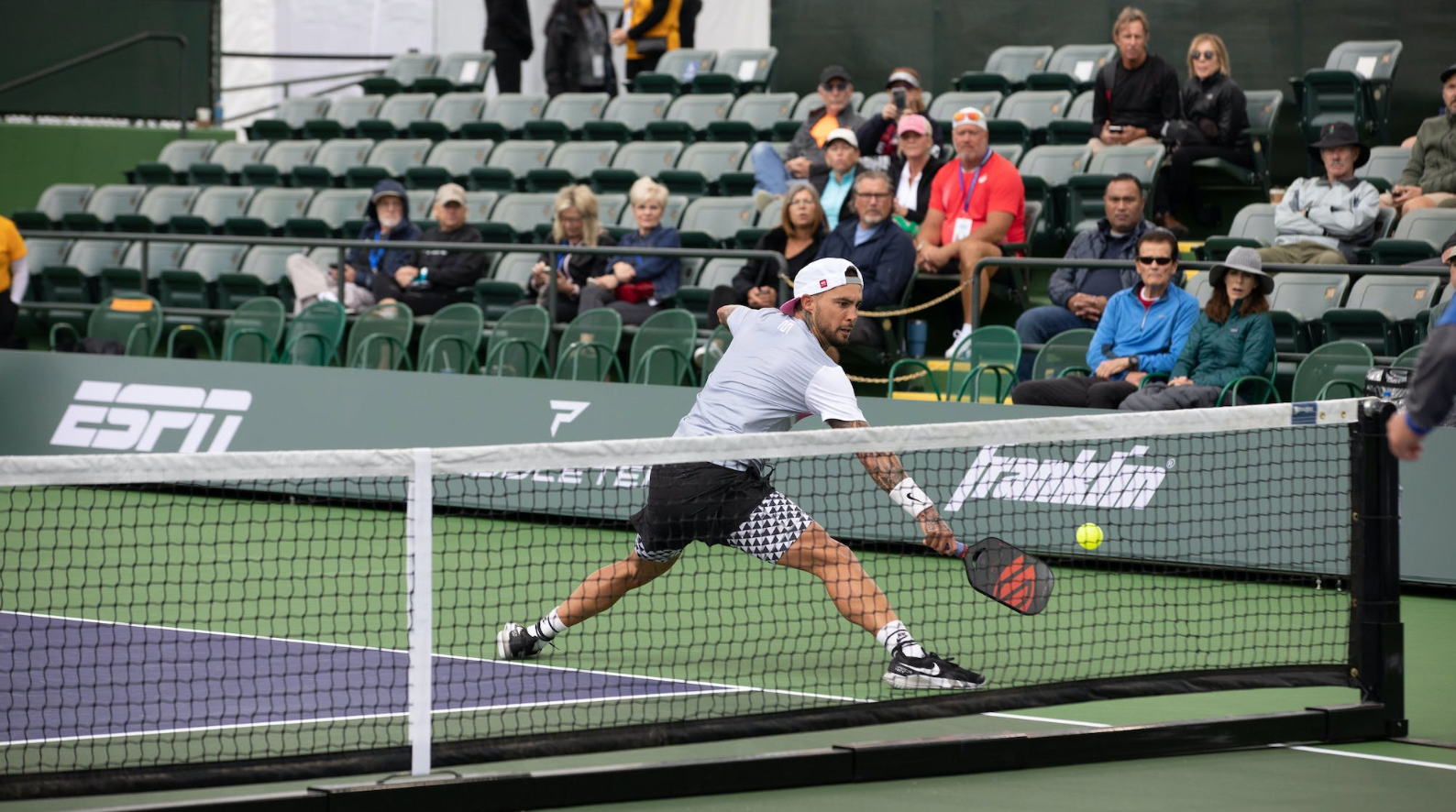 Pickleball player lunging to hit a backhand volley at the net during a match with spectators in green stadium seats