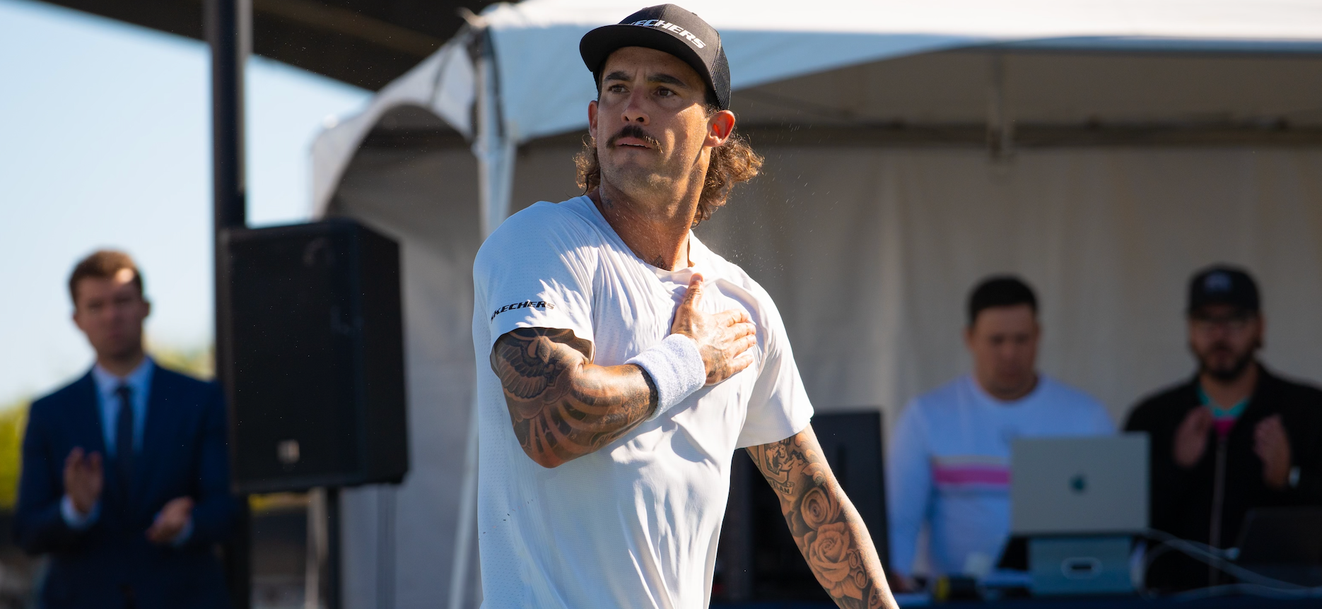 Pickleball player celebrating on outdoor stage with hand on chest, tattooed arm visible and people clapping in background
