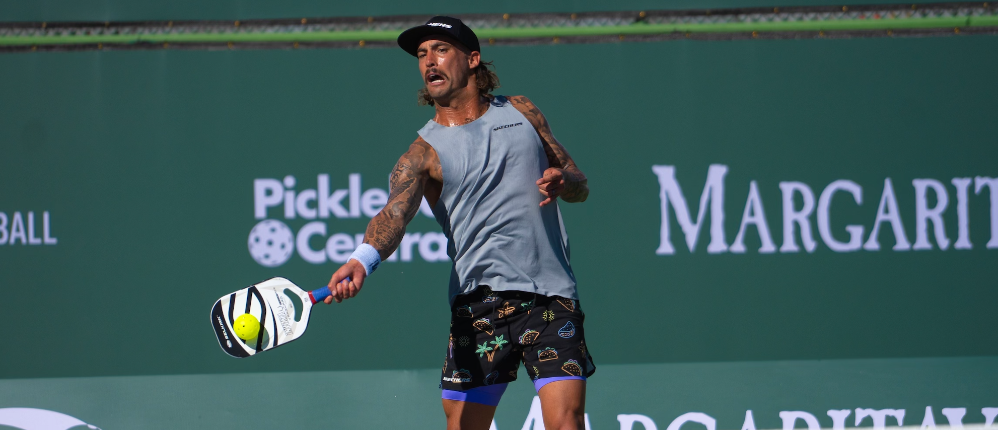 A male pickleball player in action, wearing a gray tank top and patterned shorts, preparing to hit a ball.