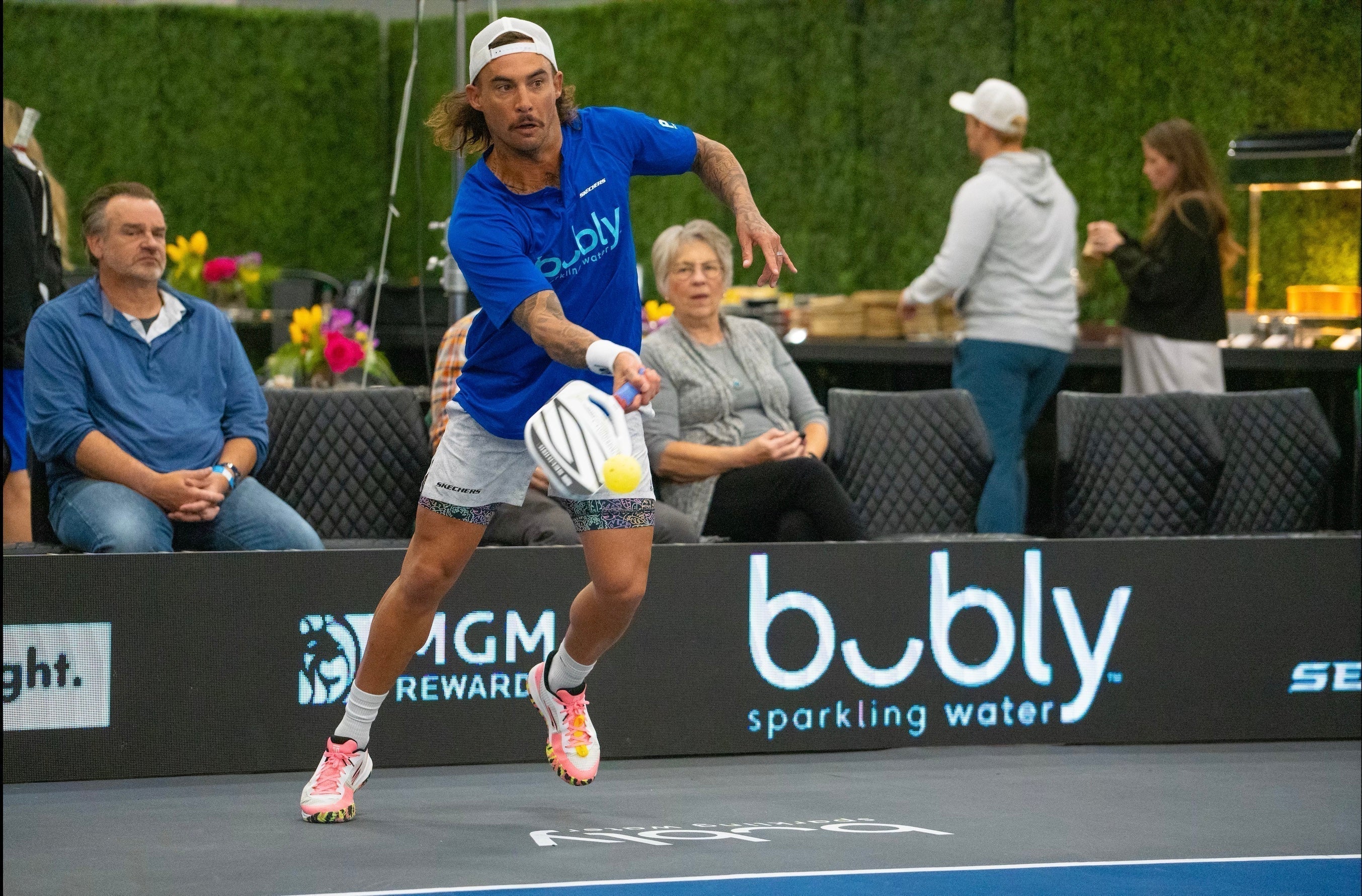A player in a blue shirt hitting a pickleball with 'bubly sparkling water' logo visible.