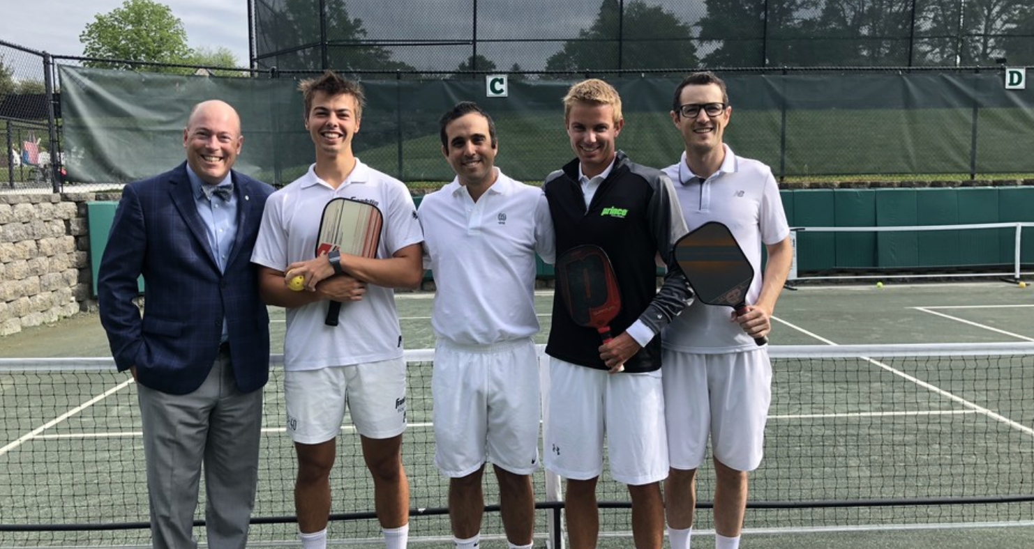 Four men in white attire holding pickleball paddles on a court, with a man in a suit beside them.