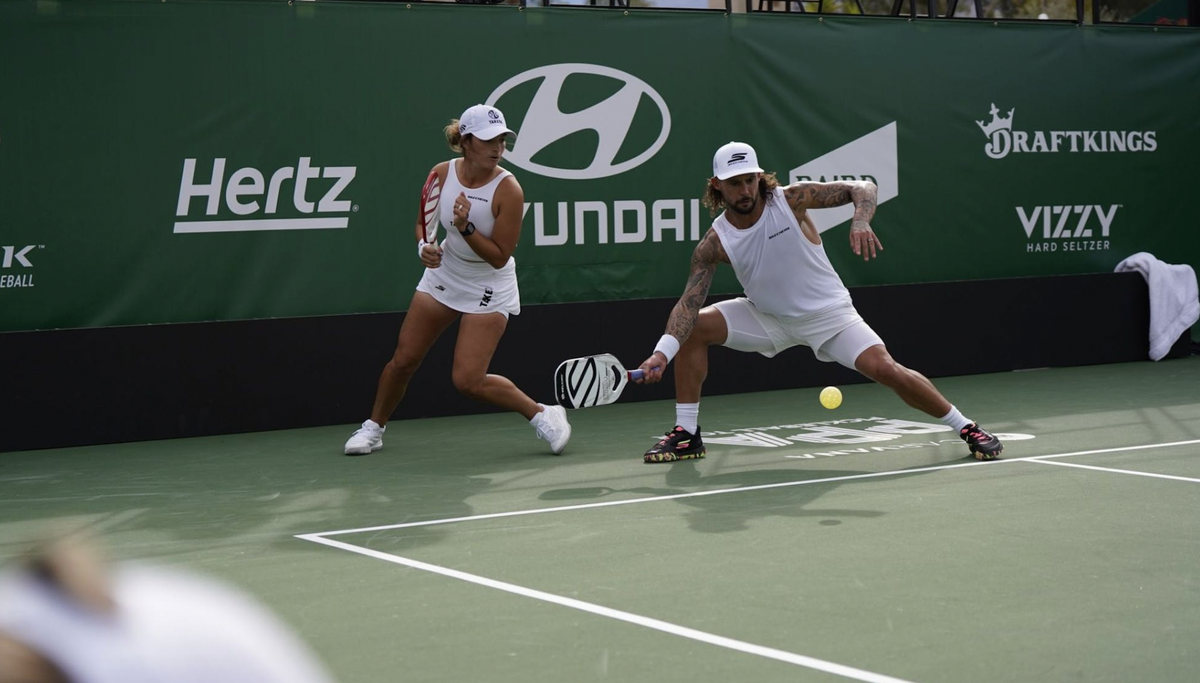 Two pickleball players lunge toward a ball on a sponsored pickleball court