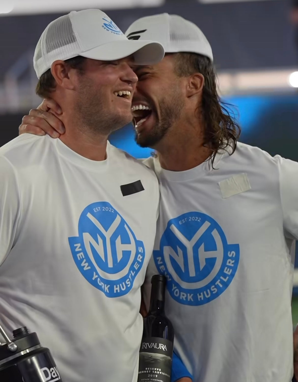 Two players from the New York Hustlers celebrating with a bottle of wine, wearing team shirts.