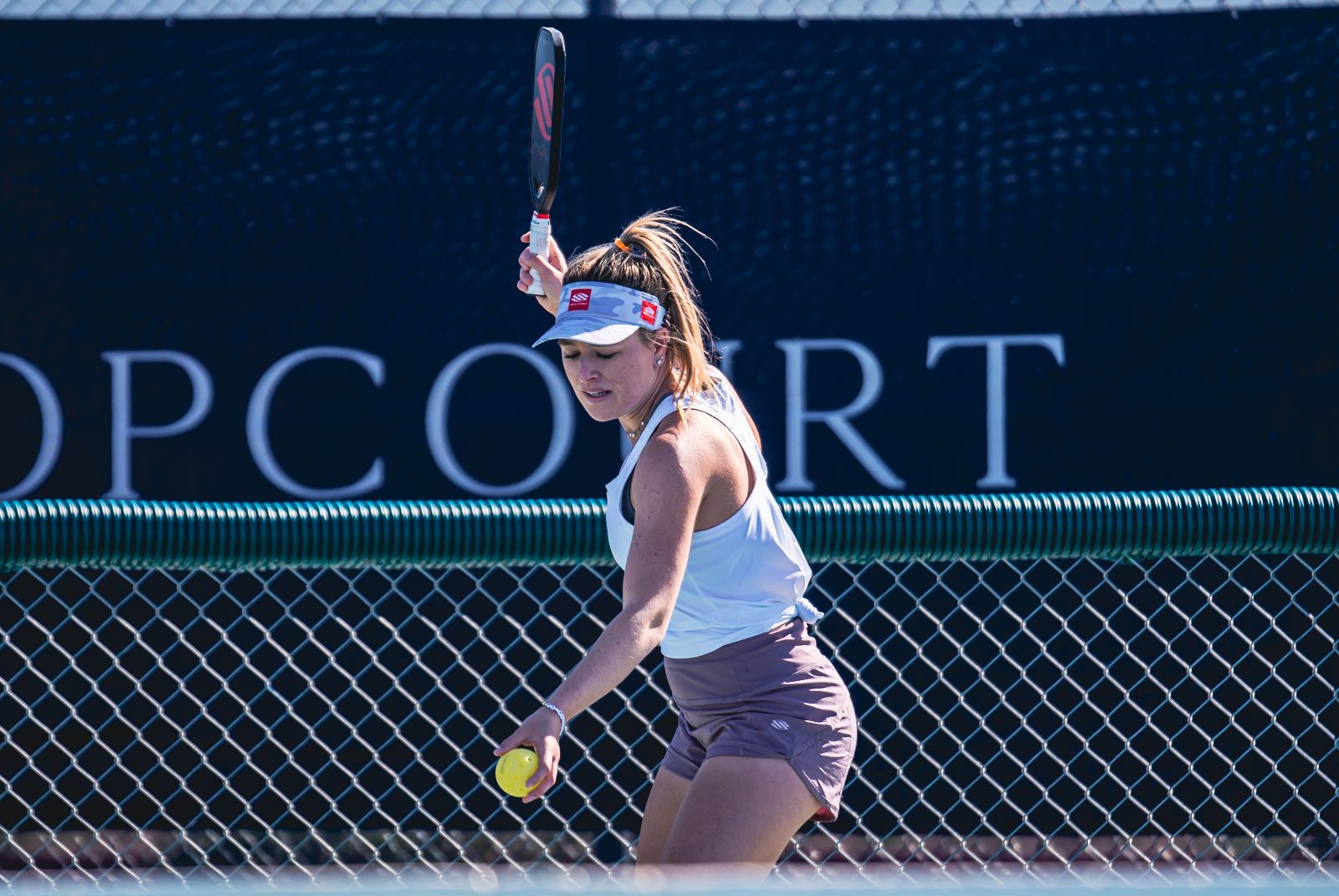 A female pickleball player in a white tank top and brown shorts prepares to hit a pickleball with a paddle at a tournament.