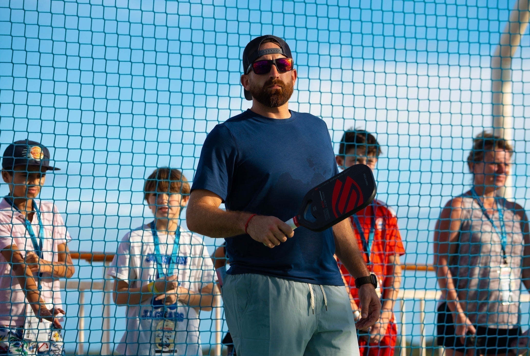 A man holding a pickleball paddle stands in front of a net with children watching.