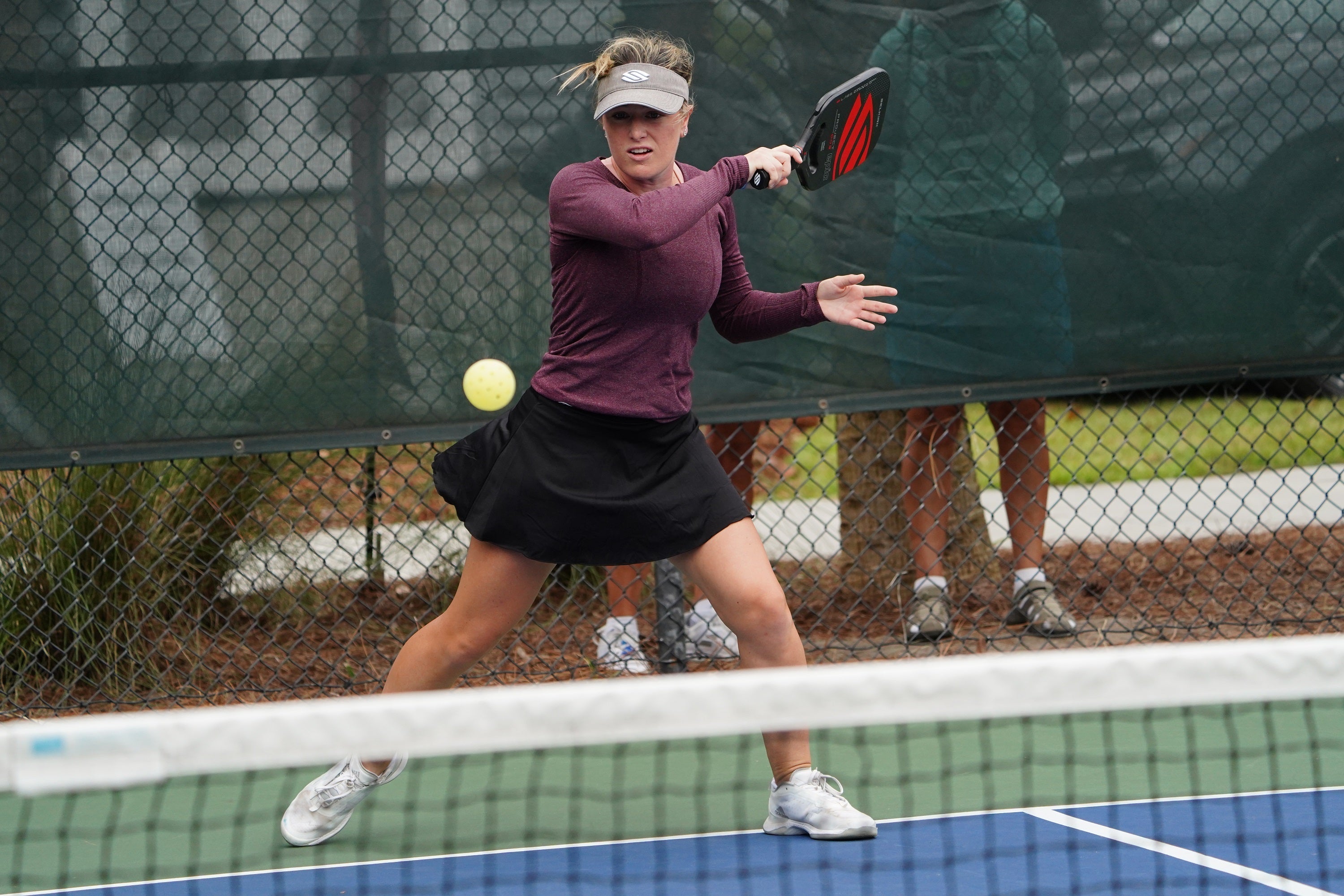 A female player in a purple long-sleeve shirt and black skirt swings a pickleball paddle at a ball on a court.