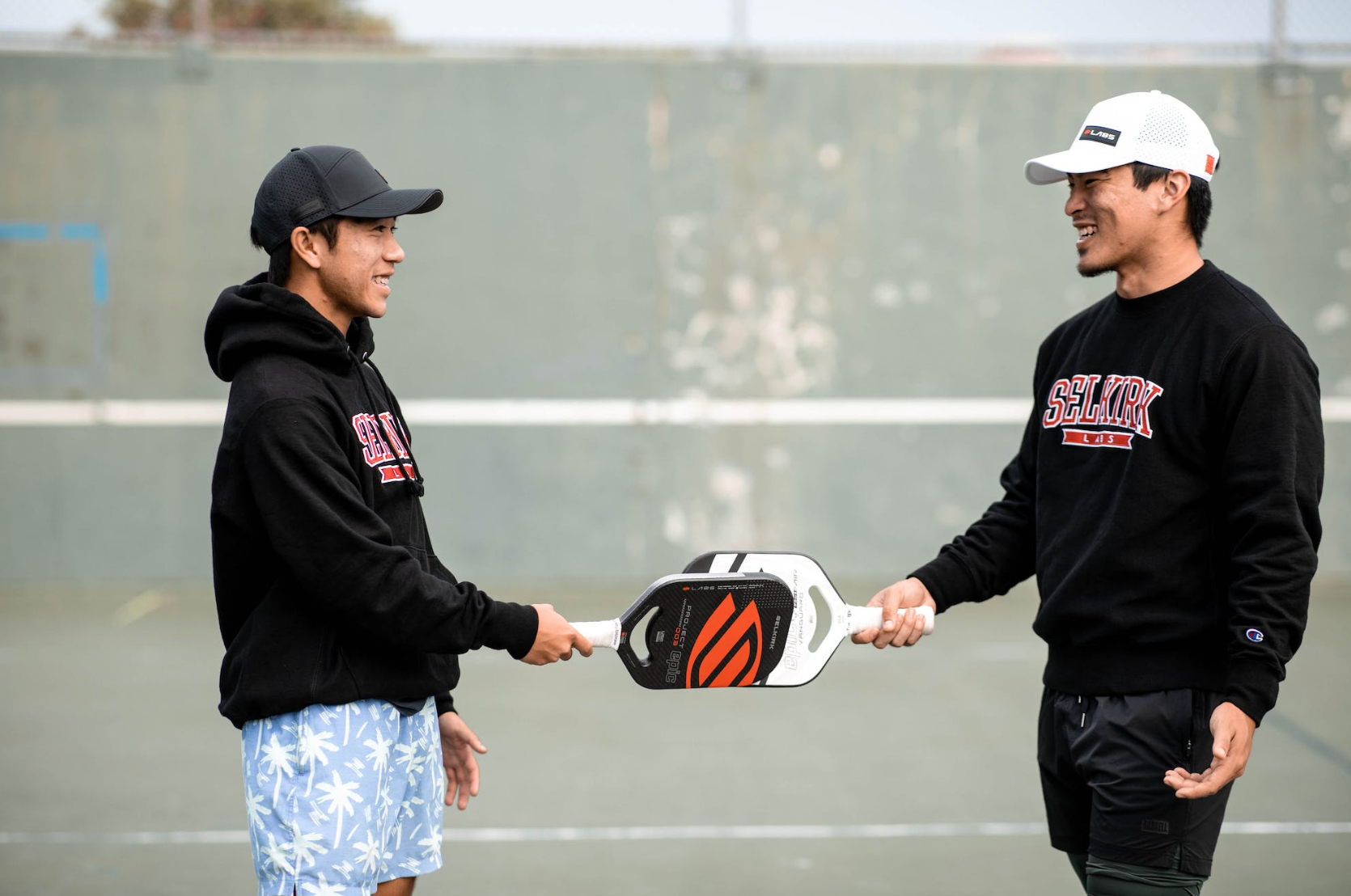 Two individuals exchanging a Selkirk pickleball paddle on a court.