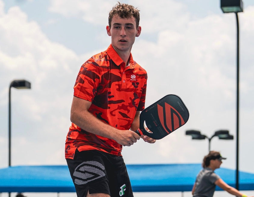 A young male pickleball player in a red camo shirt, holding a paddle, ready to play.