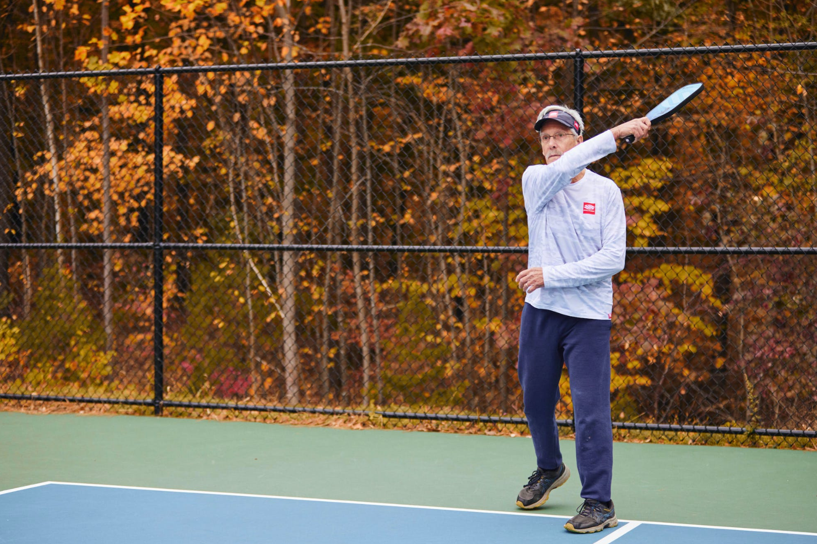 Man playing pickleball on an outdoor court with autumn trees and a chain-link fence in the background, wearing a white long-sleeve shirt, navy pants, and a visor, holding a paddle mid-swing.