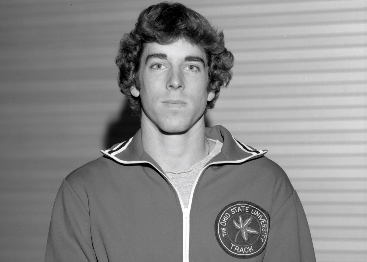 Young man facing the camera wearing a vintage zip-up athletic track jacket against a neutral ribbed background