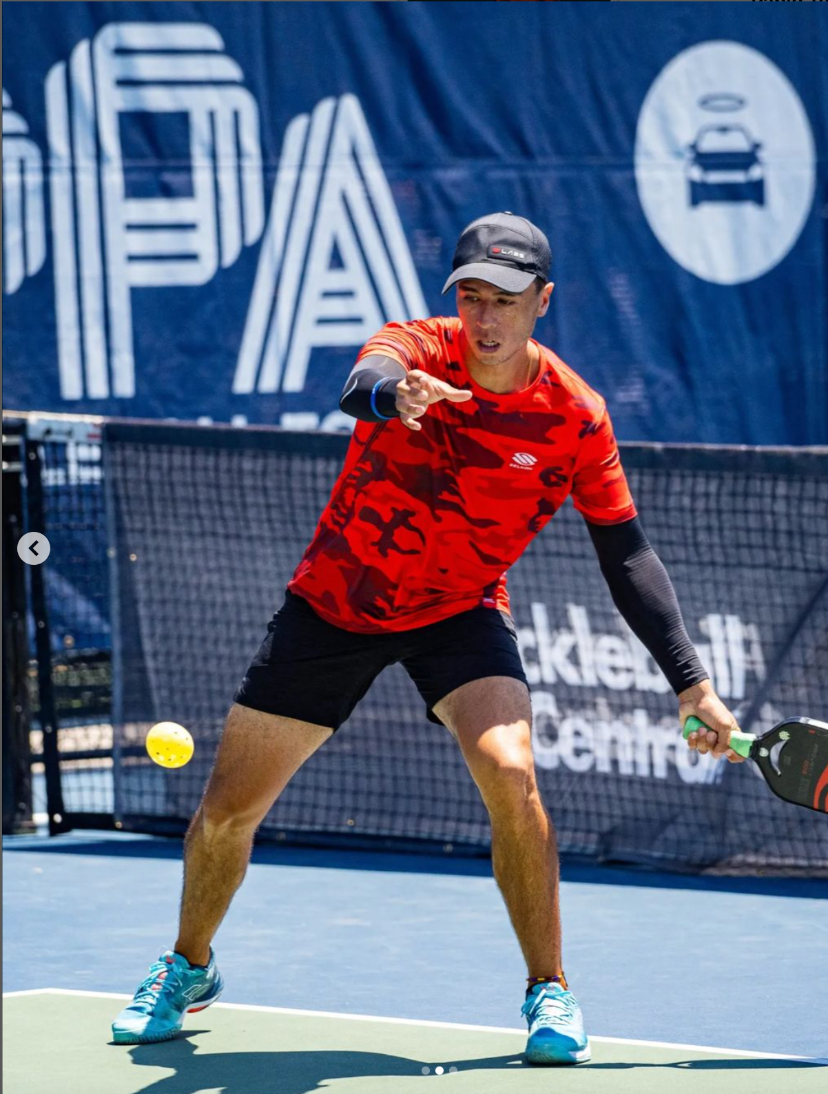 A male pickleball player in a red shirt and black shorts hitting a ball during a match.