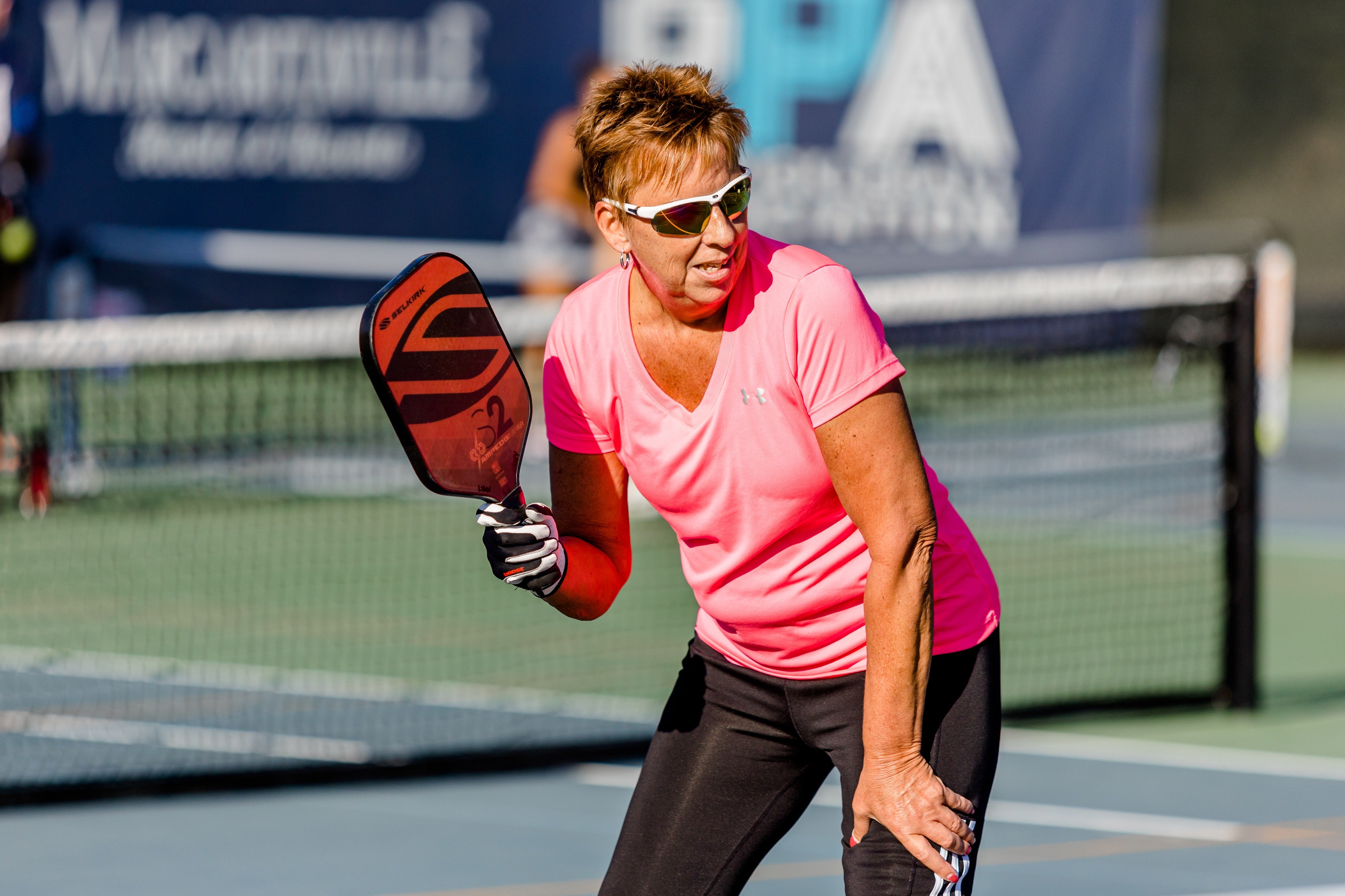 A woman in a pink shirt and sunglasses preparing to hit a pickleball with a paddle on a court.