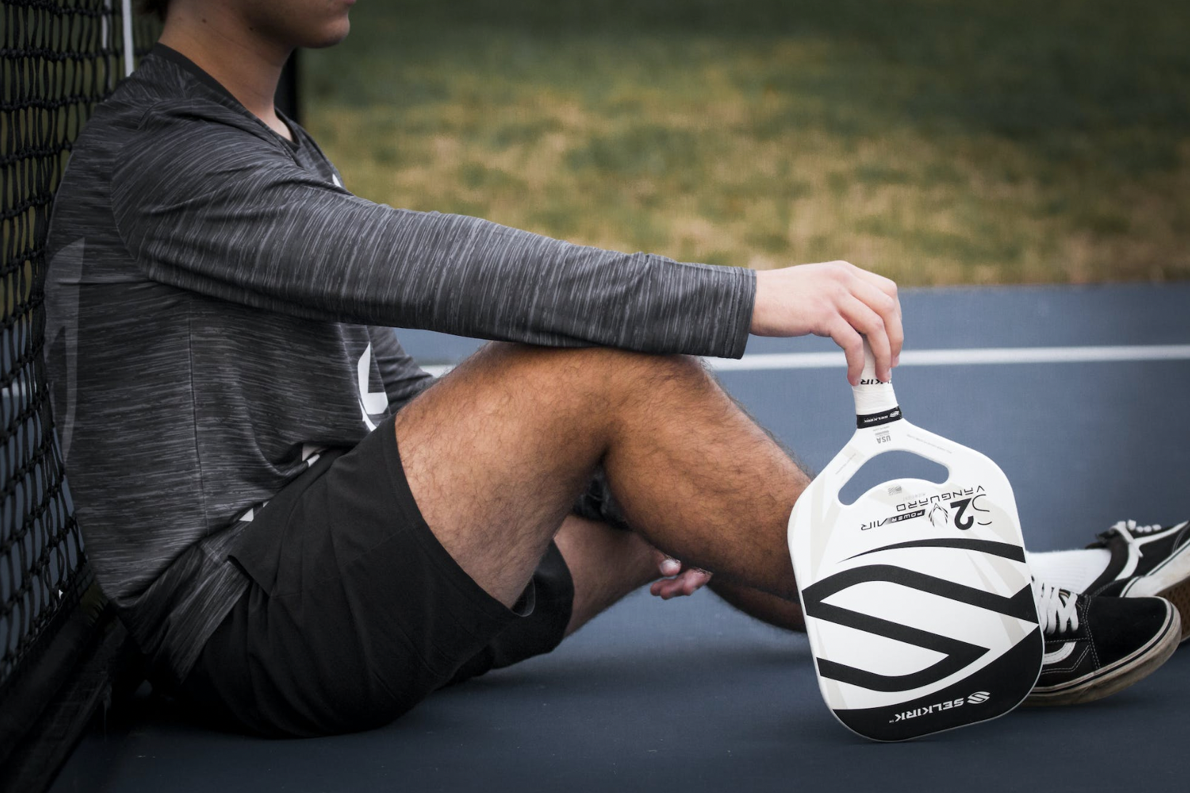 A person sitting on a court holding a pickleball paddle resting on their leg.