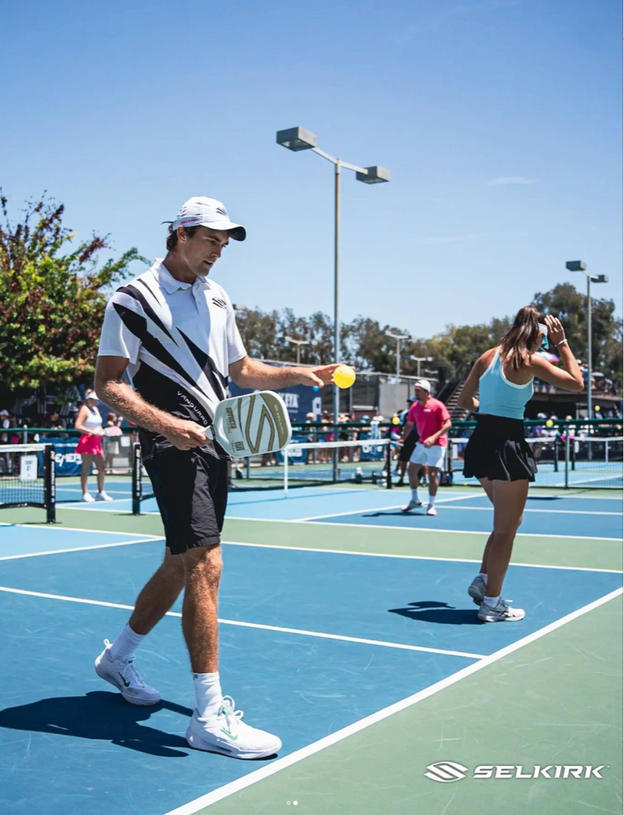 A male player in a black and white outfit prepares to serve while a female player in a skirt stands nearby on a pickleball court.