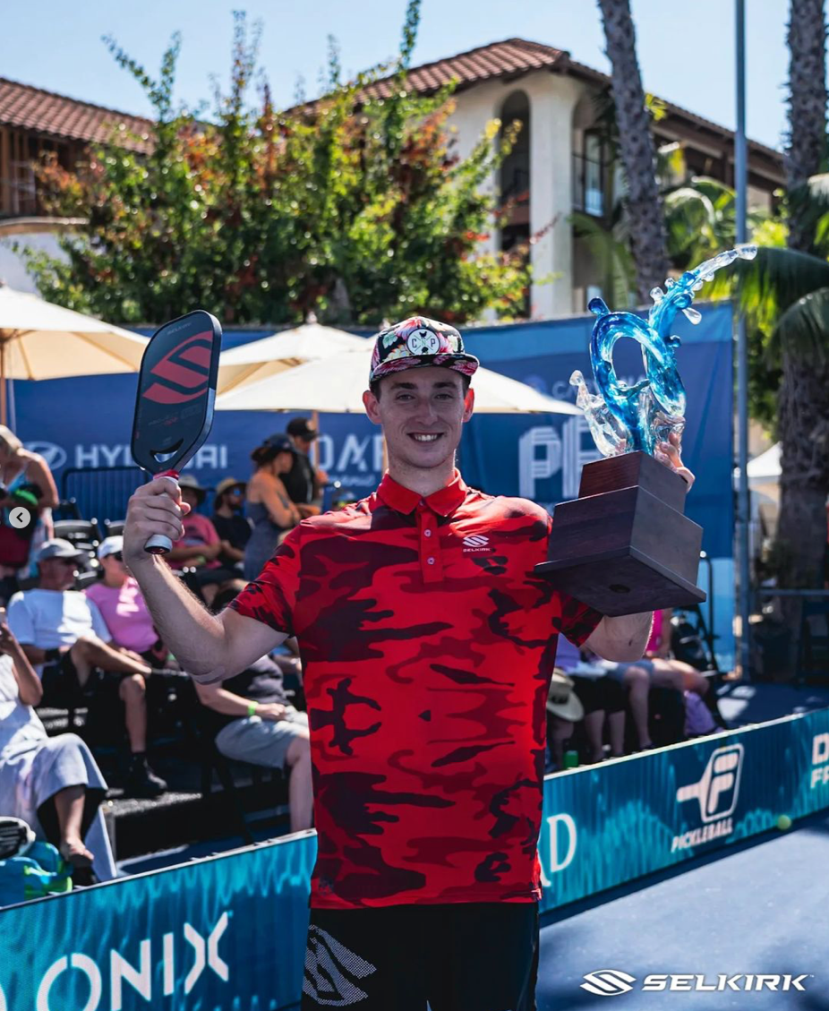 A player in a red camo shirt holding a trophy and paddles at the PPA Takeya Showcase.