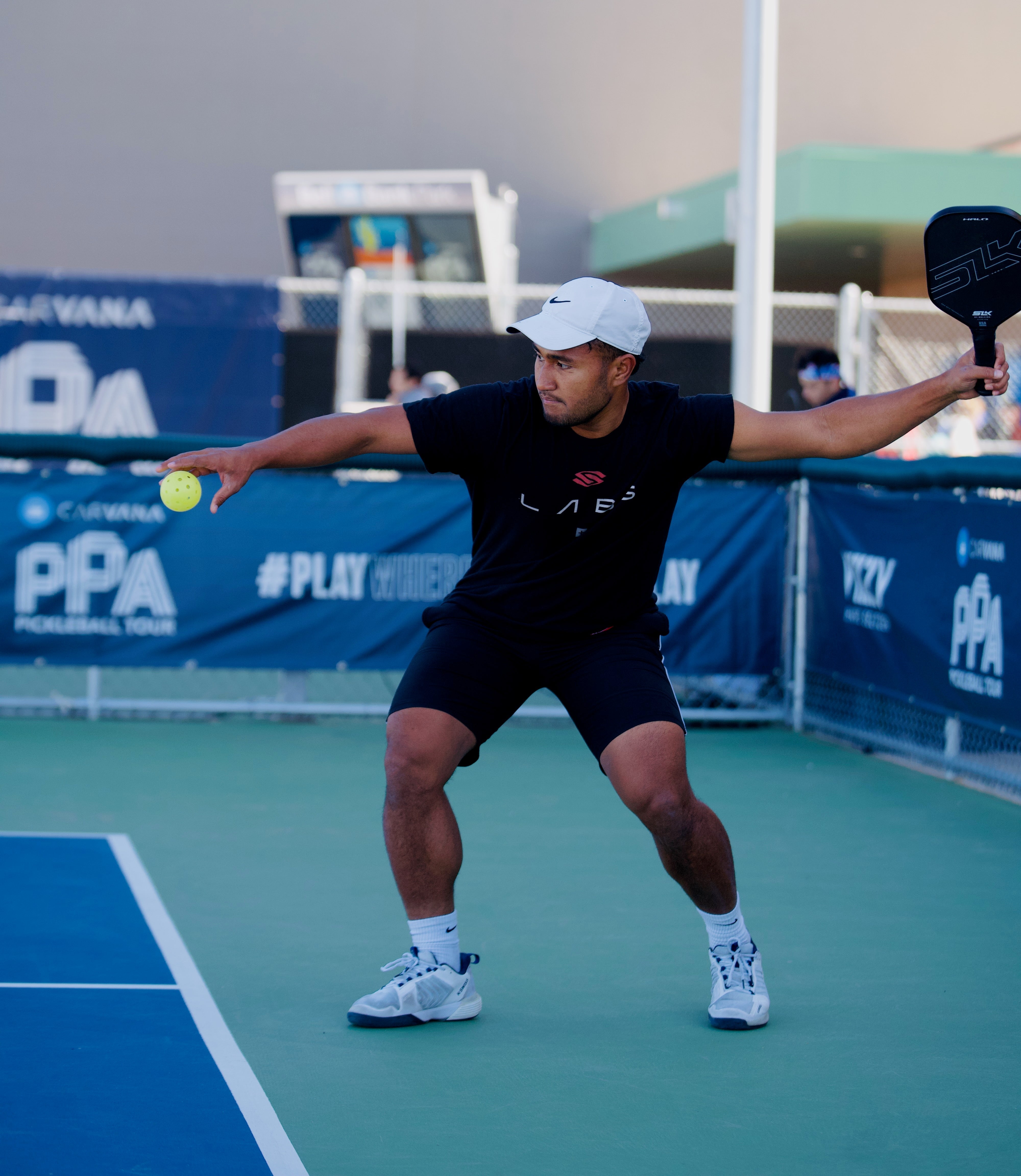 A male pickleball player serves by dropping the ball with his paddle behind him on an outdoor court.