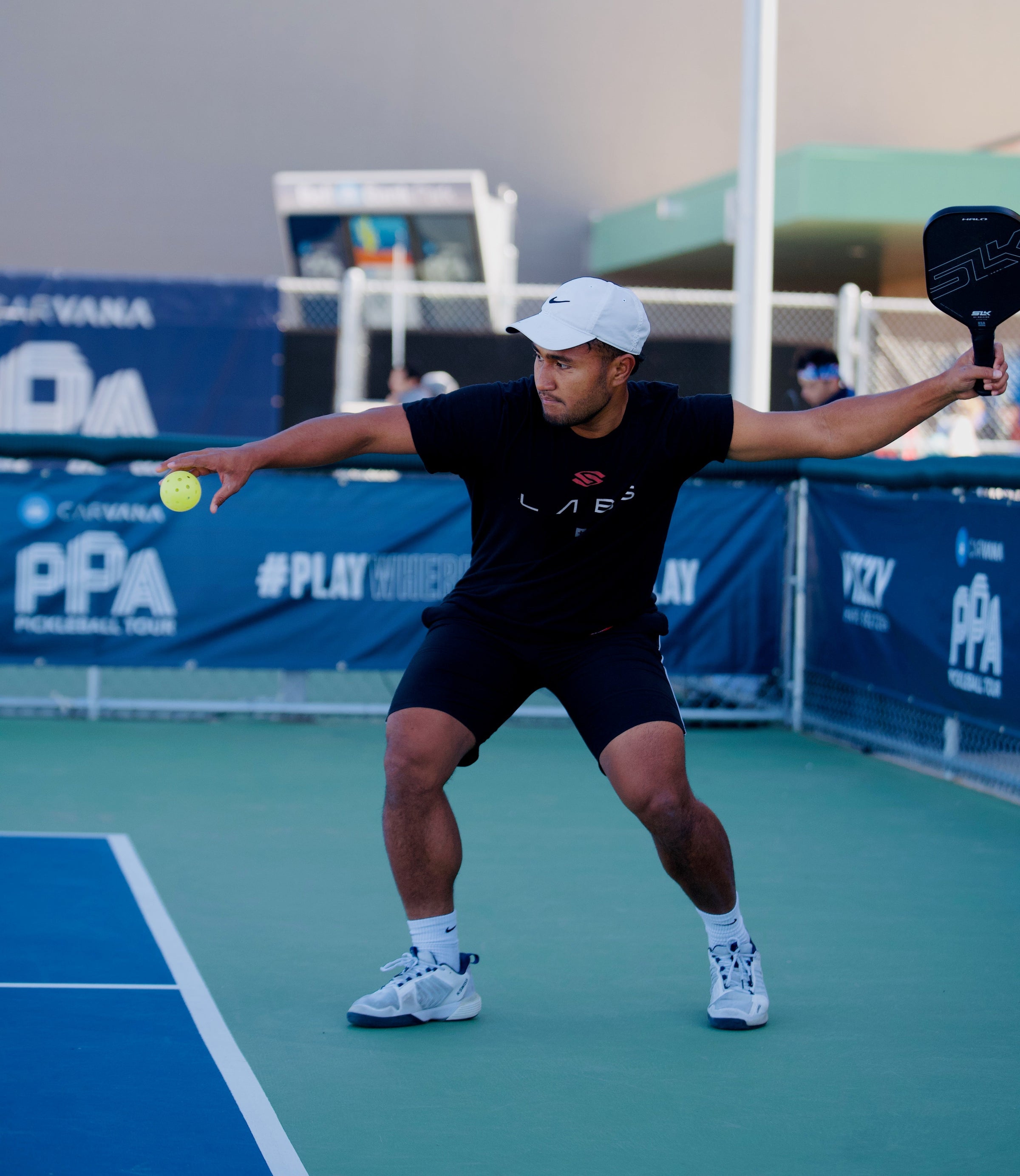 A male pickleball player serves by dropping the ball with his paddle behind him on an outdoor court.