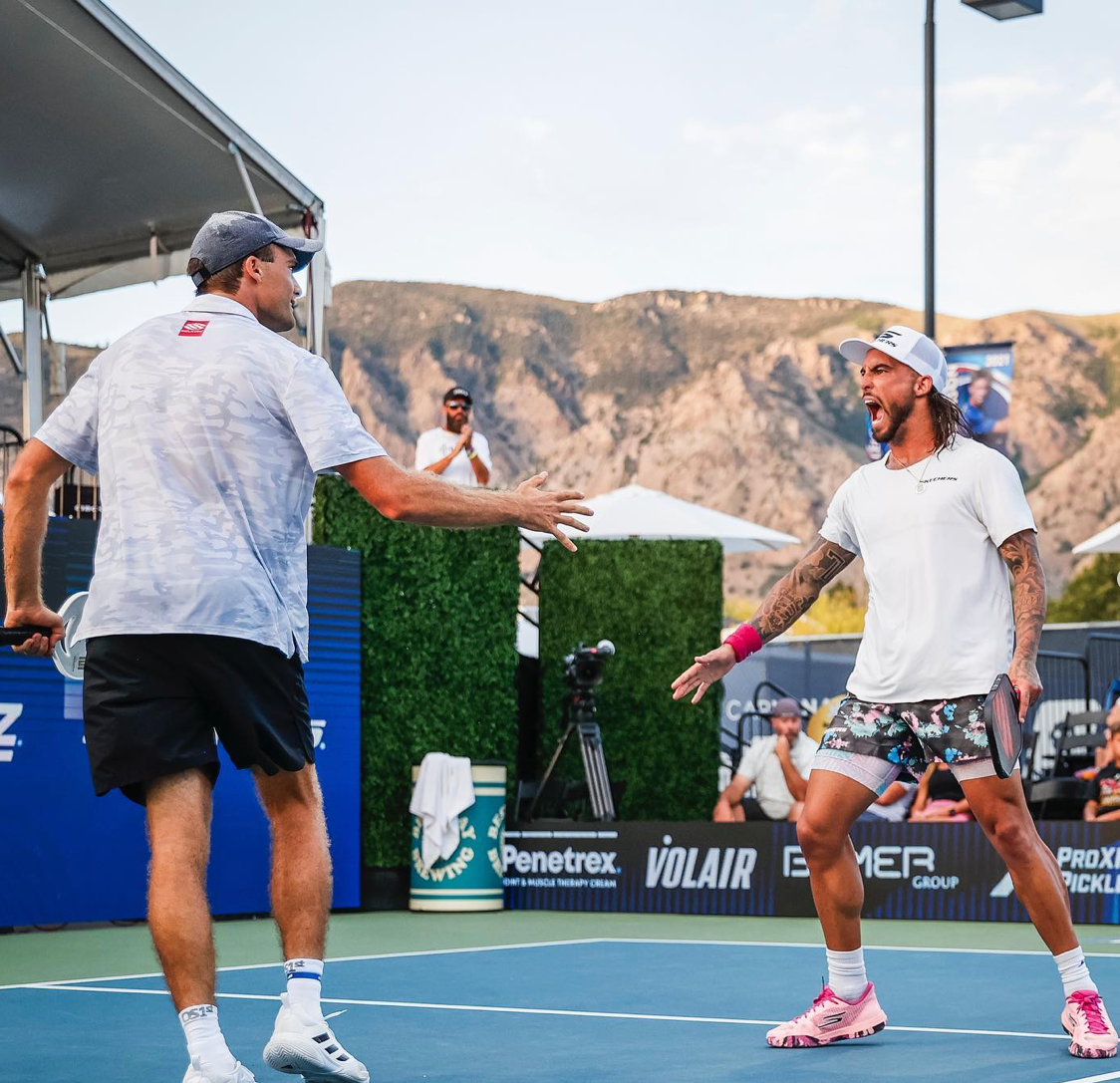 Two male pickleball players celebrating on the court after a match, with mountains in the background.