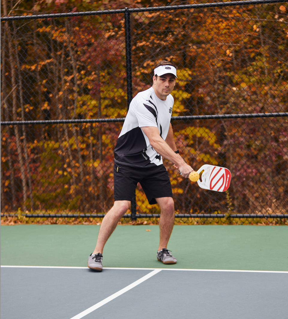 A player preparing to hit a pickleball on a court surrounded by autumn foliage.