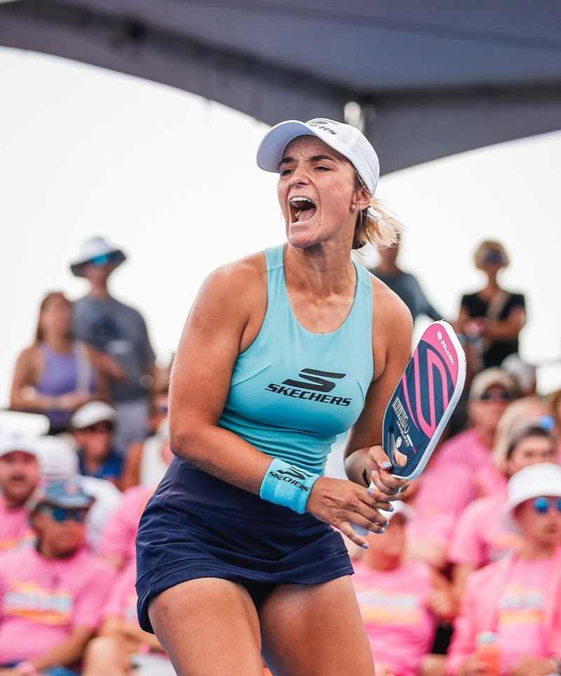A female pickleball player celebrating with a paddle, wearing a blue tank top and white cap, with spectators in the background.