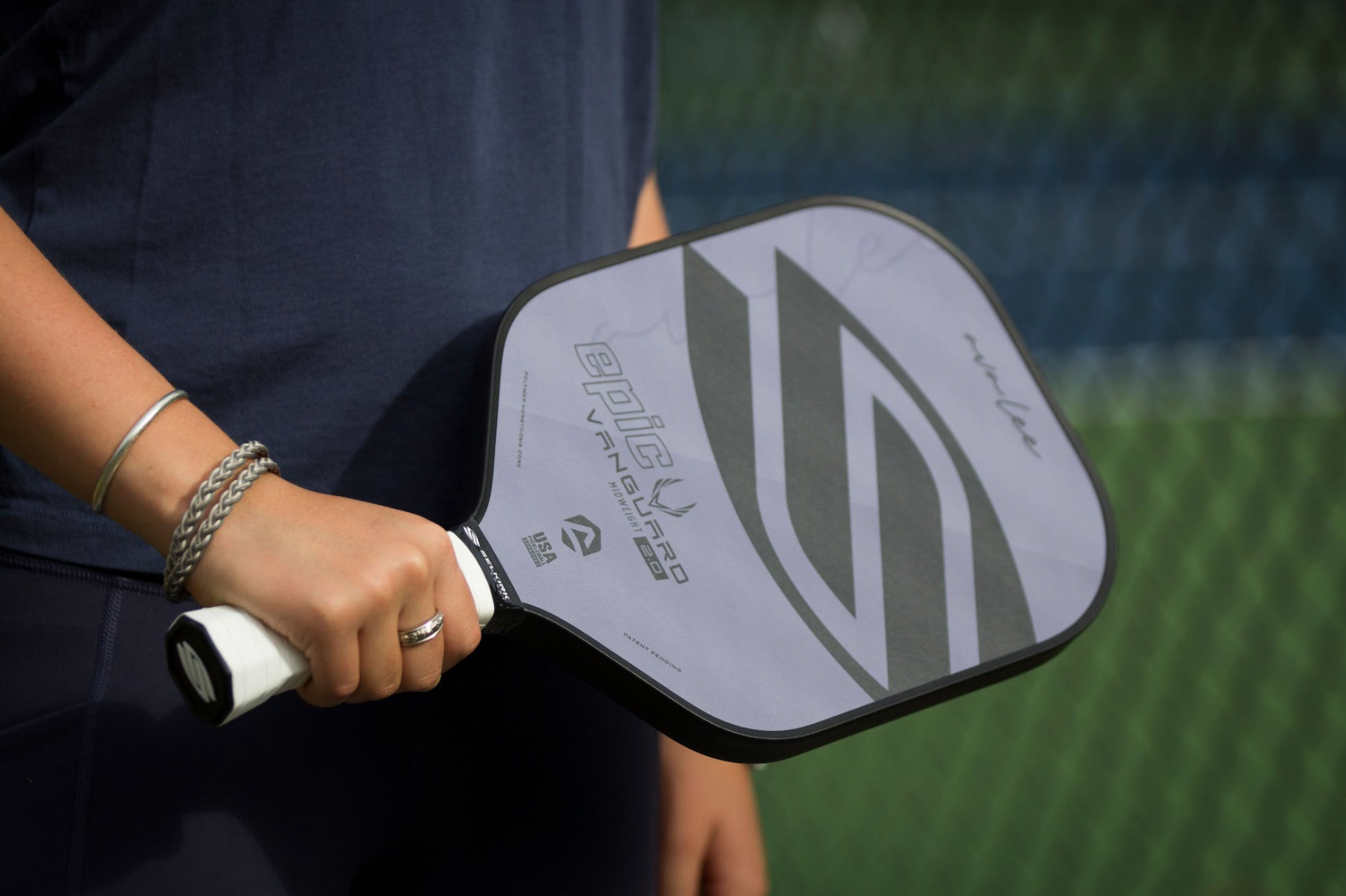 Close-up of Epic Vanguard 2.0 pickleball paddle showing textured face and black edge guard, held by a player in front of a chain-link fence